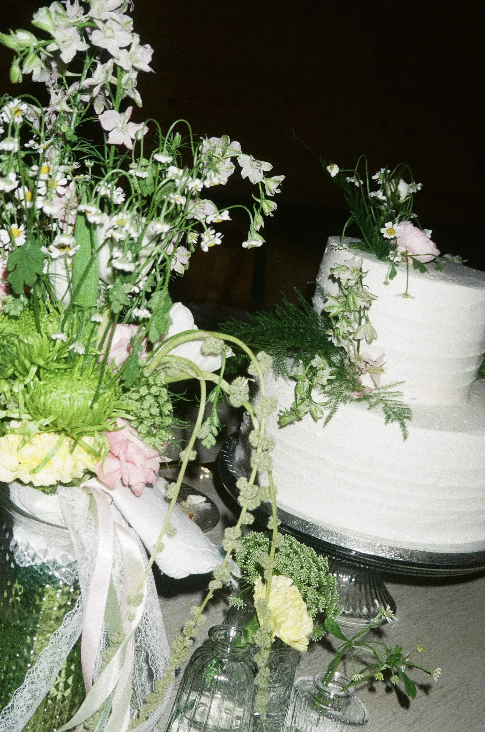 Arrangement of flowers and greenery on a table, including a white cake with floral decorations and glass jars filled with flowers, in a dark background setting.