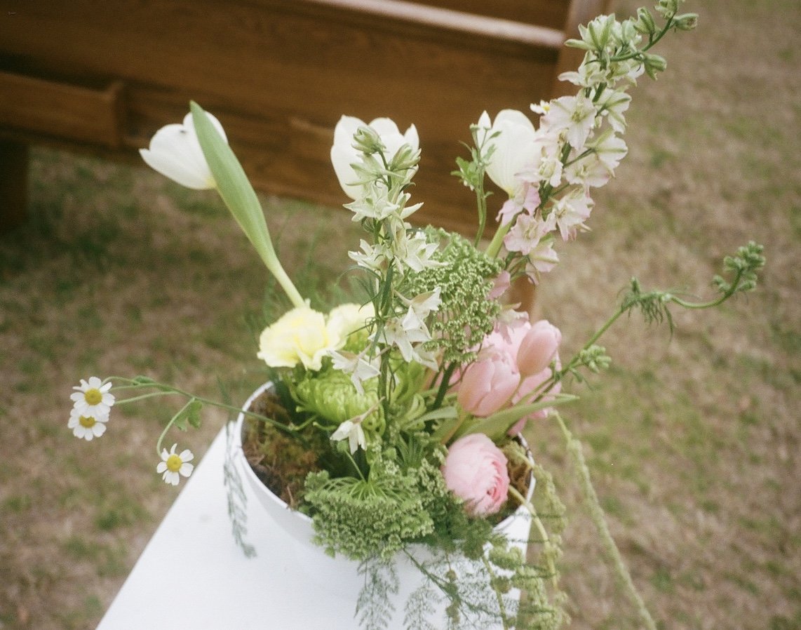 A white vase filled with various fresh flowers including daisies, tulips, and other small flowers with green foliage, placed on a white surface outdoors.