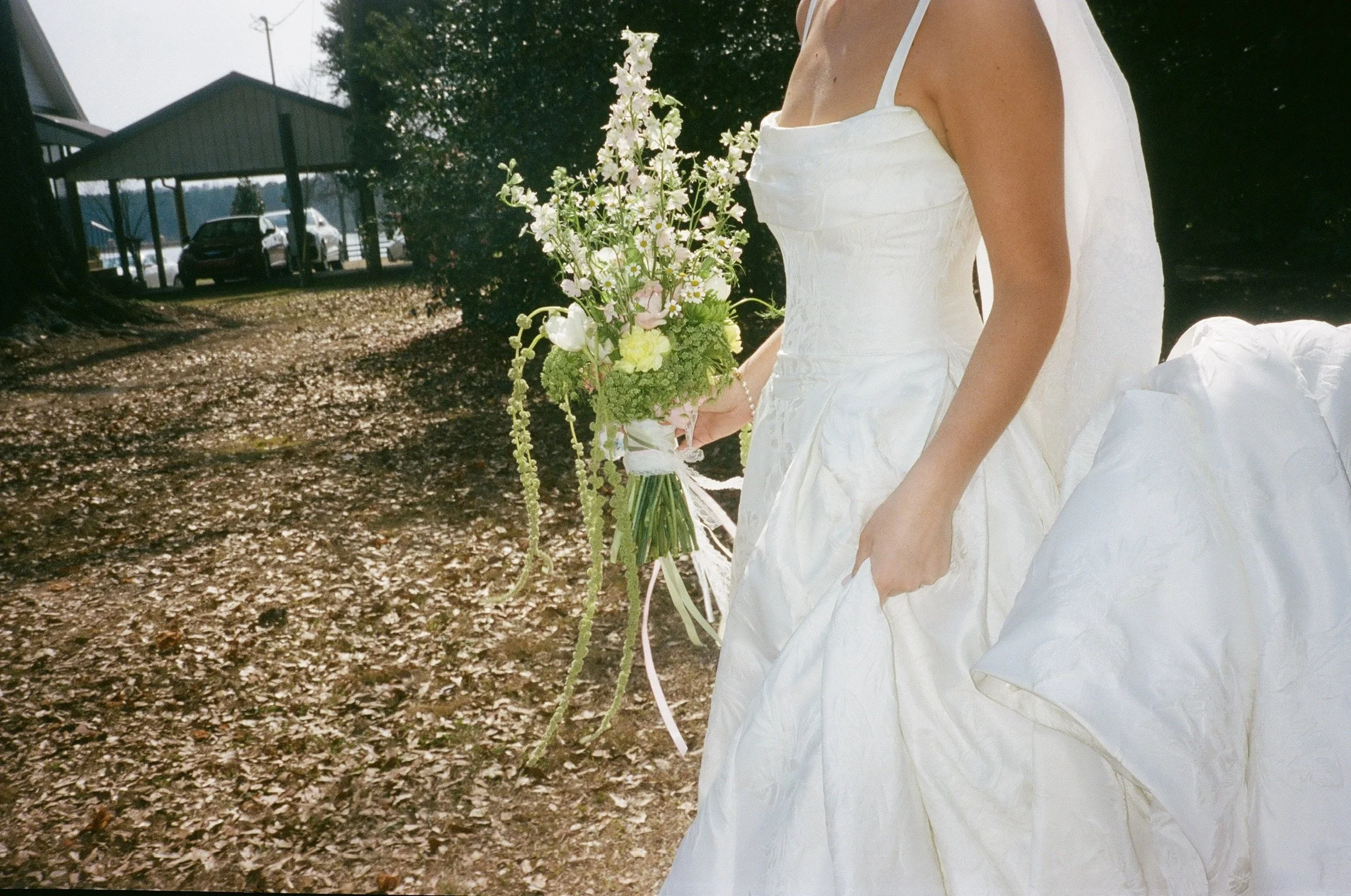 A woman in a white dress holding a bouquet of flowers, outdoors in a wooded area with a house and cars in the background.
