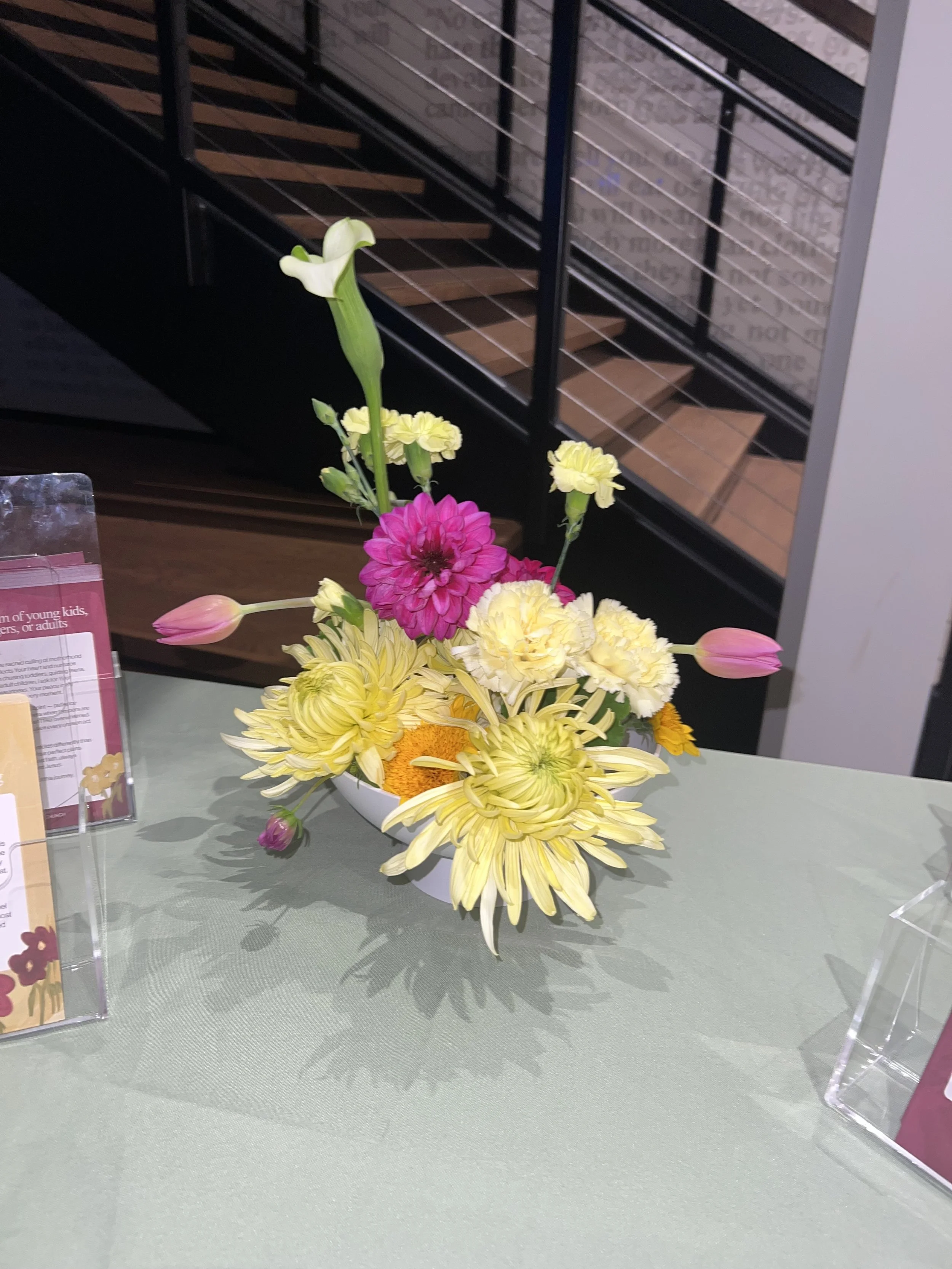 A flower arrangement in a white vase with white, pink, yellow, and purple flowers, placed on a green tablecloth, with a staircase in the background.
