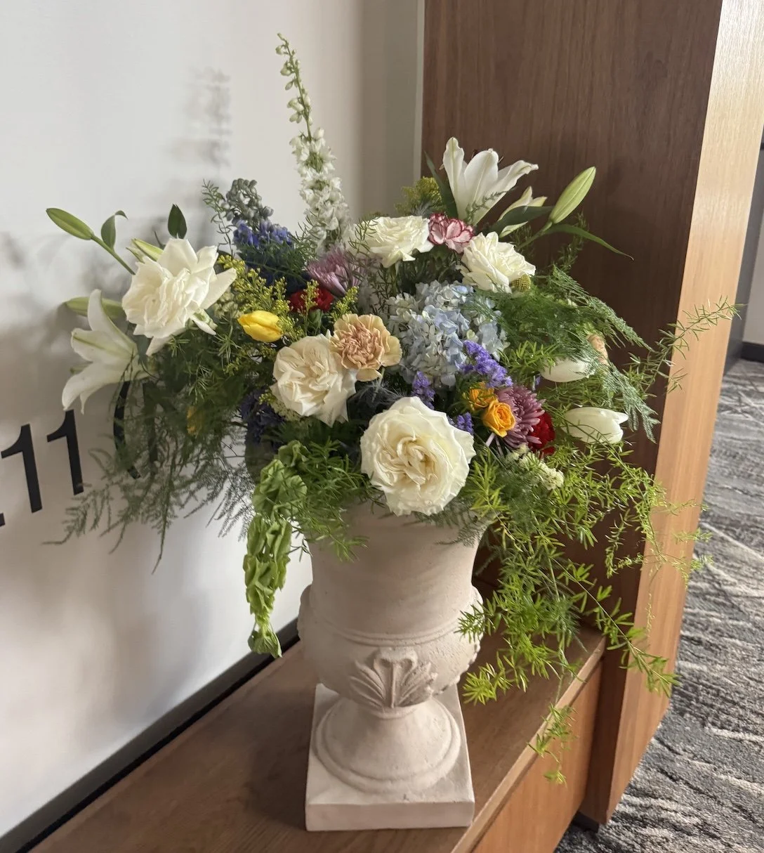 A large floral arrangement in a decorative white urn with various white, purple, yellow, red, pink, and green flowers, placed on a wooden surface next to a white wall and wooden cabinet.