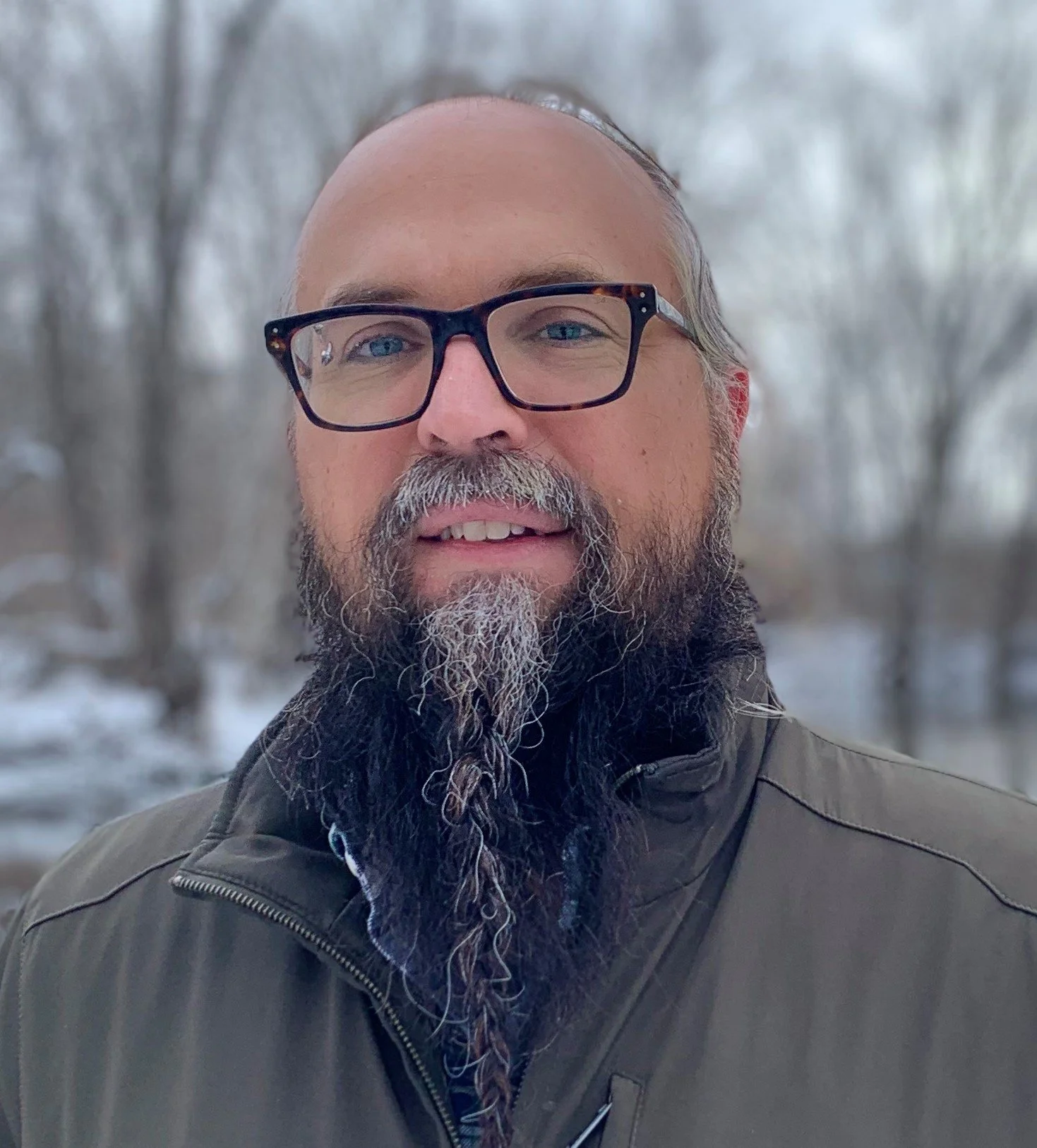 A man with glasses and a beard standing outdoors in a snowy, wooded landscape.