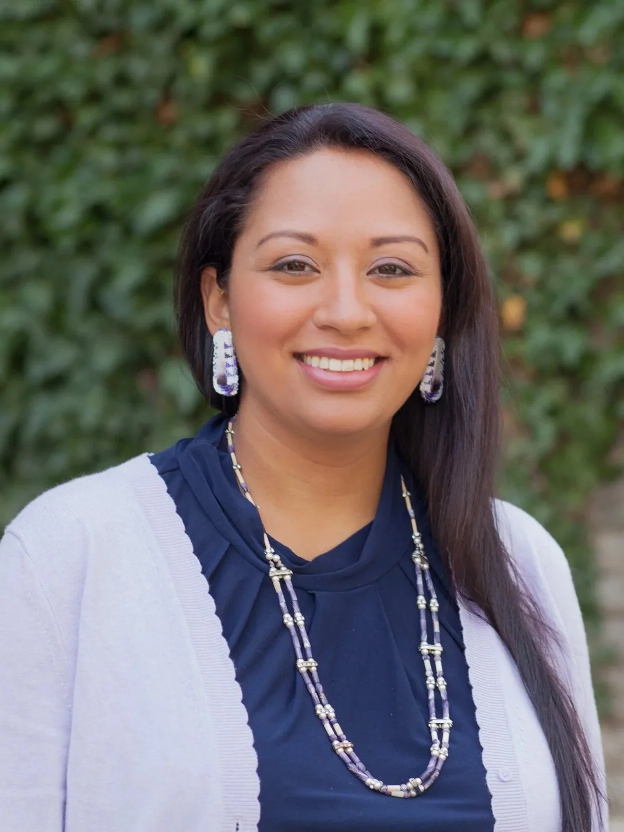 A woman with dark hair and brown eyes smiling outdoors, wearing a navy blue top, white cardigan, and jewelry including earrings and layered necklaces, with a blurred green leafy background.