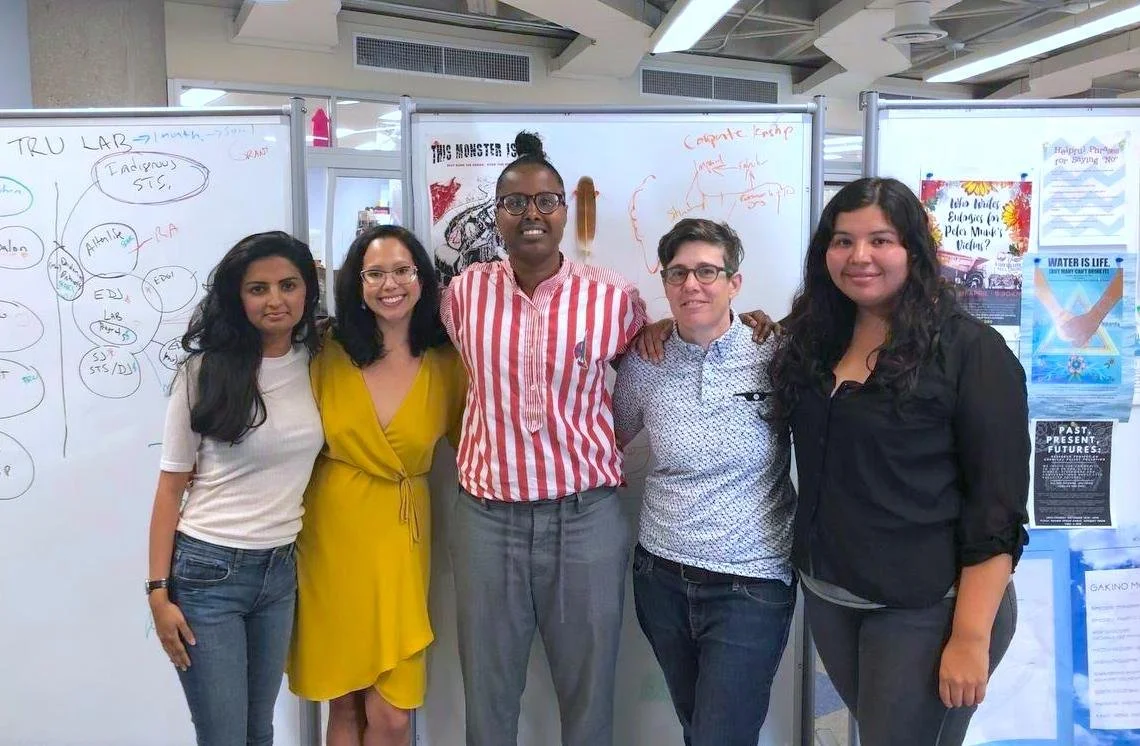 Group of five people standing in a classroom or conference room, smiling, with whiteboards and posters behind them.
