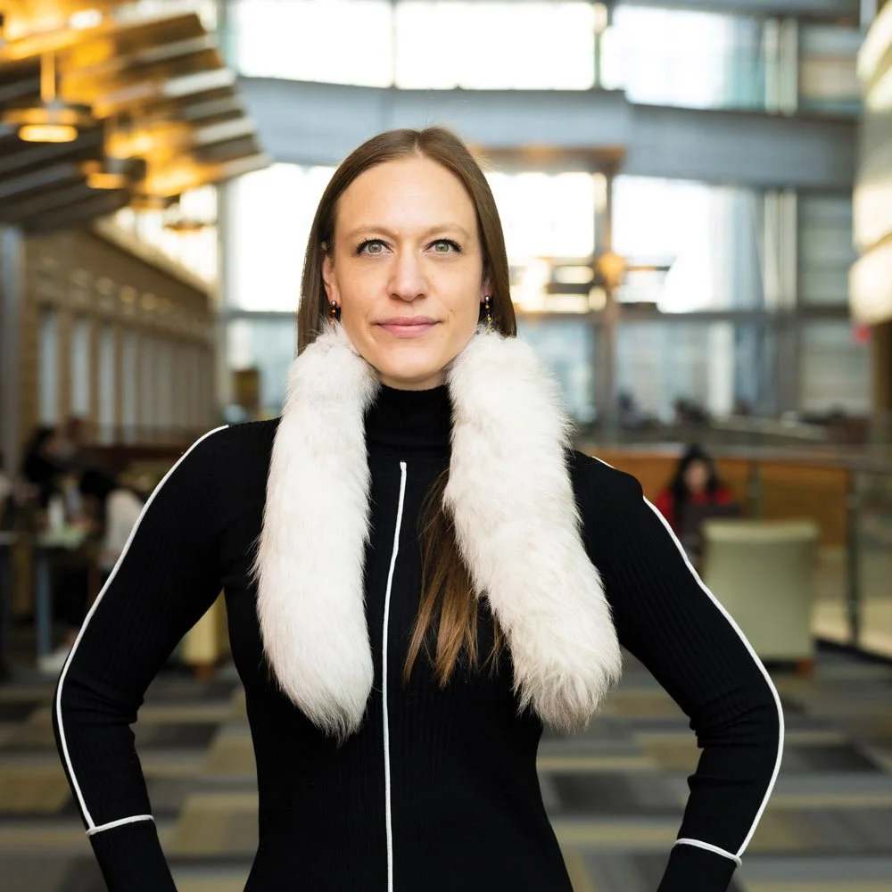 A woman with long brown hair, wearing a black top with white fur scarf, standing in an indoor space with high ceilings and large windows.