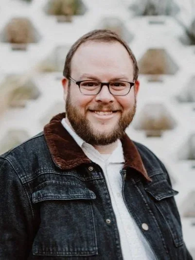 A man with glasses and a beard smiling, wearing a dark denim jacket and a white shirt, standing outdoors with a decorative brick wall in the background.