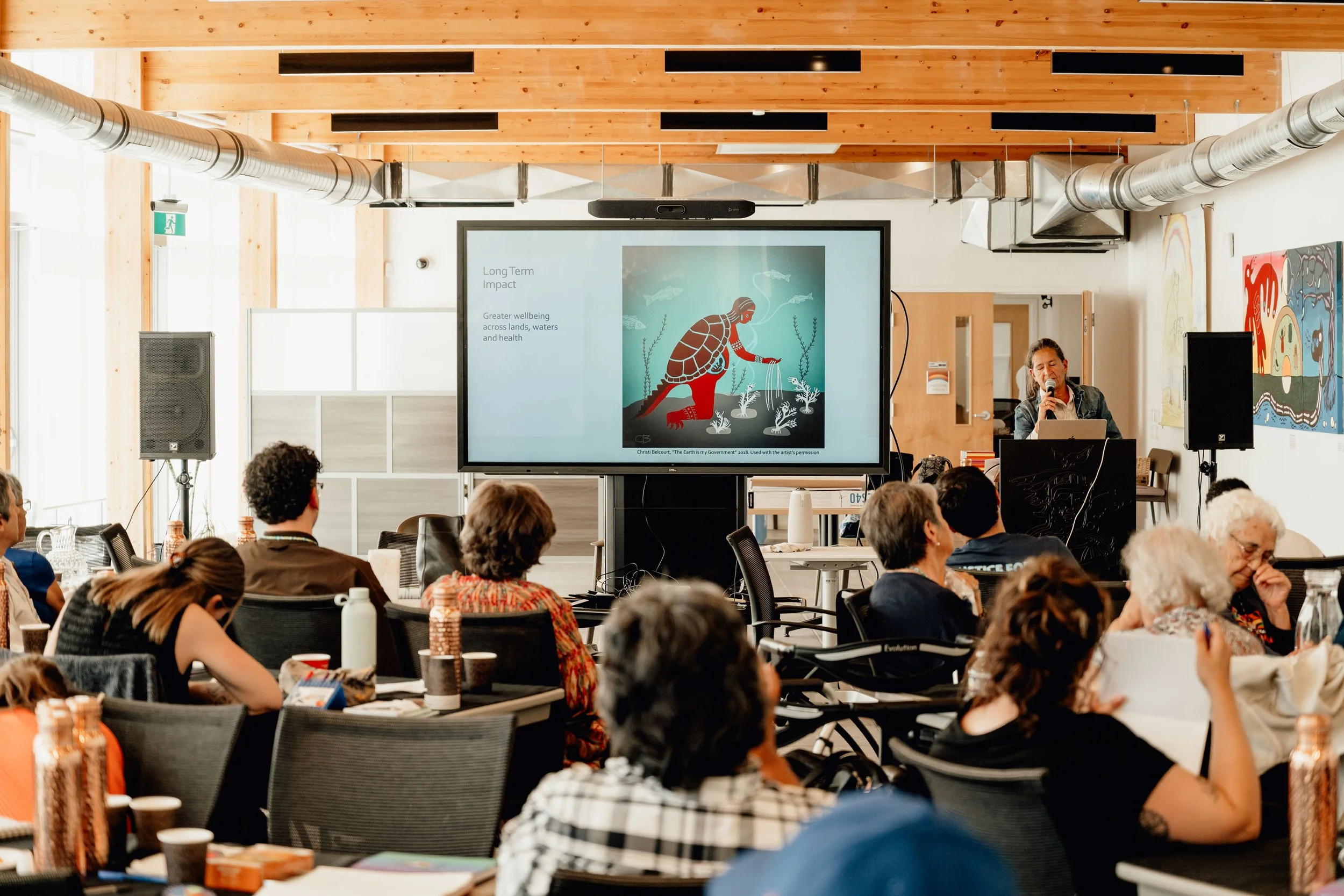 A conference or seminar held in a spacious, well-lit room with attendees seated at tables. A presenter is speaking into a microphone near a large screen displaying a slide about environmental impacts with an illustration of a turtle and fishes.