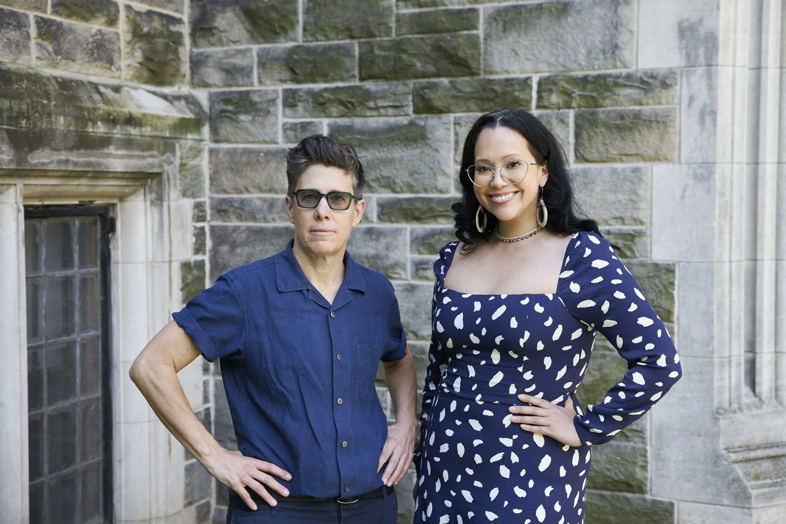 Two women standing outdoors in front of a stone building, posing confidently for the photo.