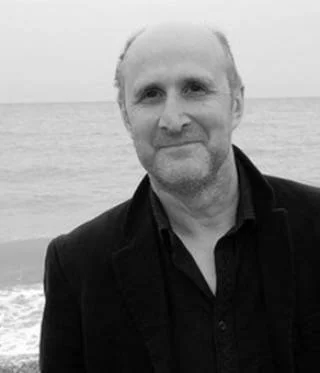 A middle-aged man with short hair and a beard, wearing a dark jacket, standing outdoors near the ocean with waves in the background.