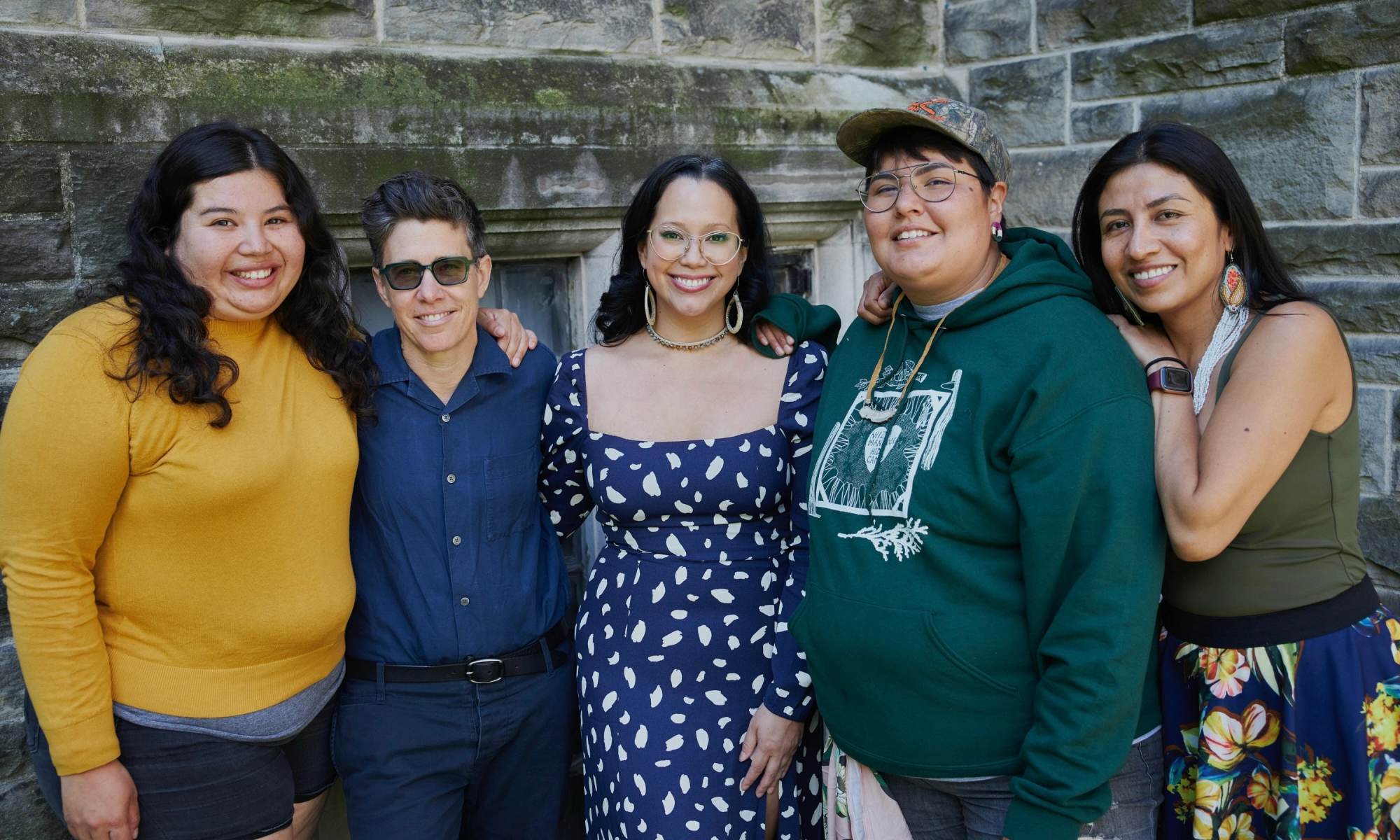Group of five diverse young adults standing outdoors in front of a stone wall, smiling at the camera.