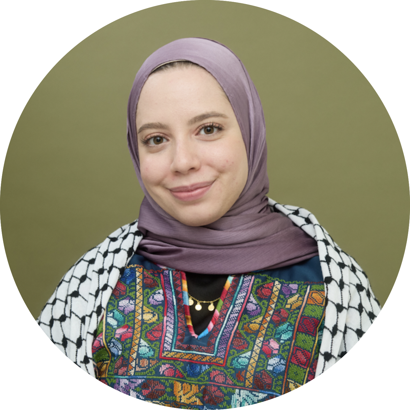 A young woman wearing a lavender hijab, traditional colorful embroidered attire, and a black and white patterned kuffiyeh, smiling and standing against a green background.