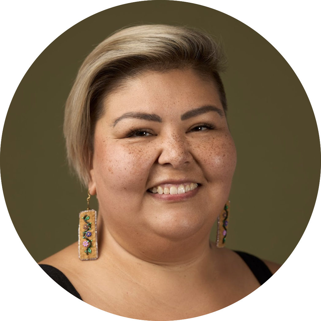 Close-up portrait of a smiling woman with short blonde hair, freckles, and colorful beaded earrings, against a plain olive green background.