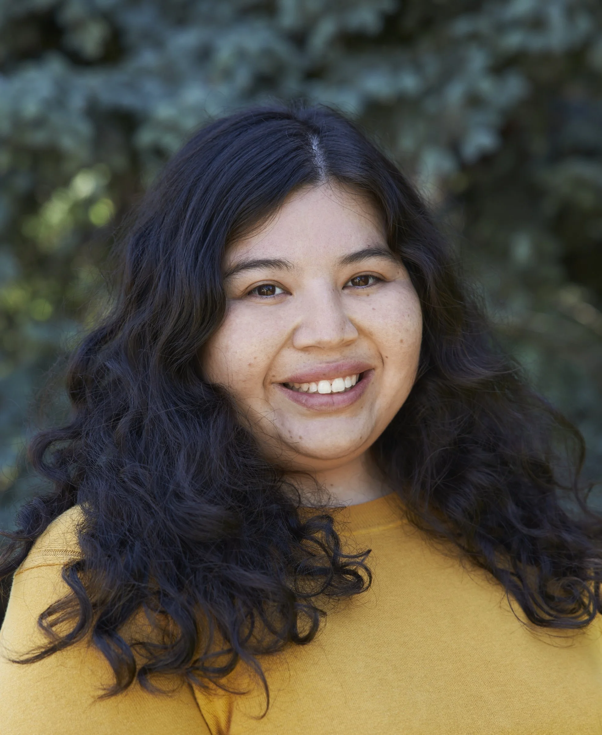 Close-up of a woman with dark, curly hair wearing a mustard-colored top, smiling outdoors with green foliage in the background.