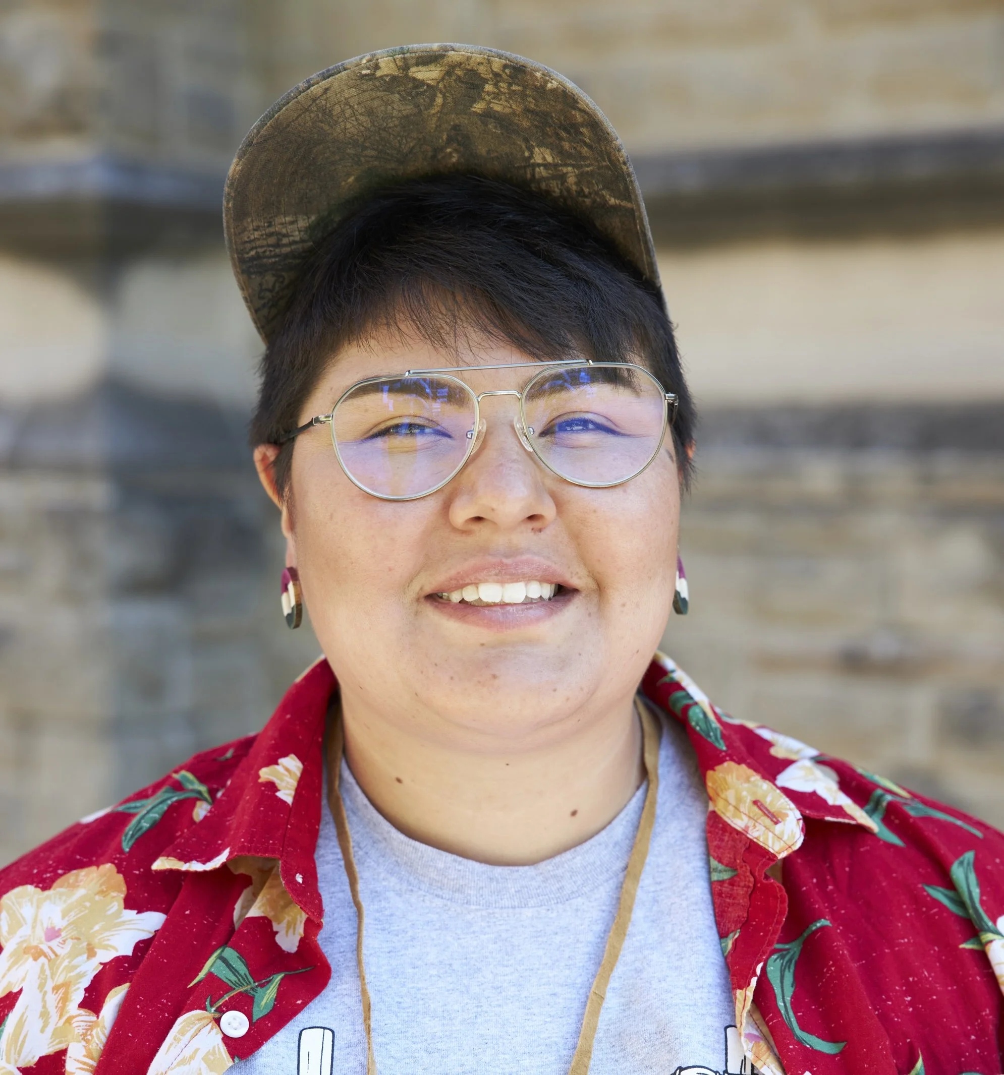 Close-up of a person wearing glasses and a red Hawaiian shirt with floral patterns, smiling, with a weathered baseball cap on their head, against a blurred outdoor background.