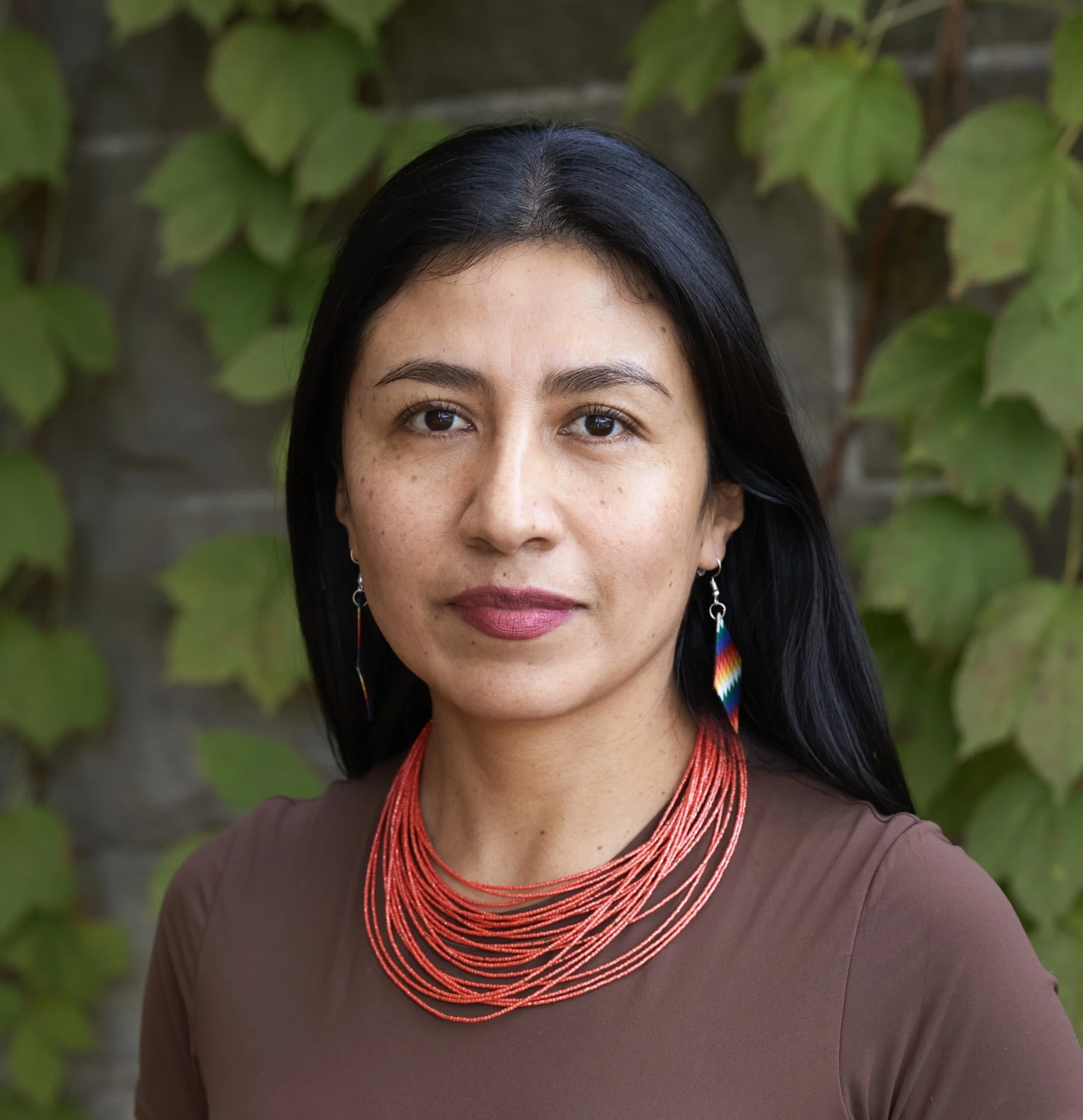 Close-up of a woman's face and shoulders with black hair, brown eyes, wearing red beaded jewelry and colorful earrings, against a background of green leaves and vine-covered wall.