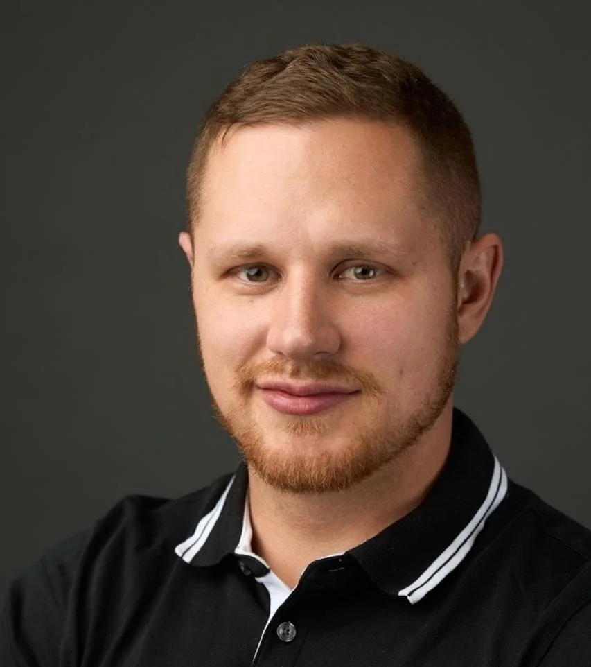 Headshot of a young man with short reddish hair and beard, wearing a black collared shirt, smiling slightly, against a plain dark background.