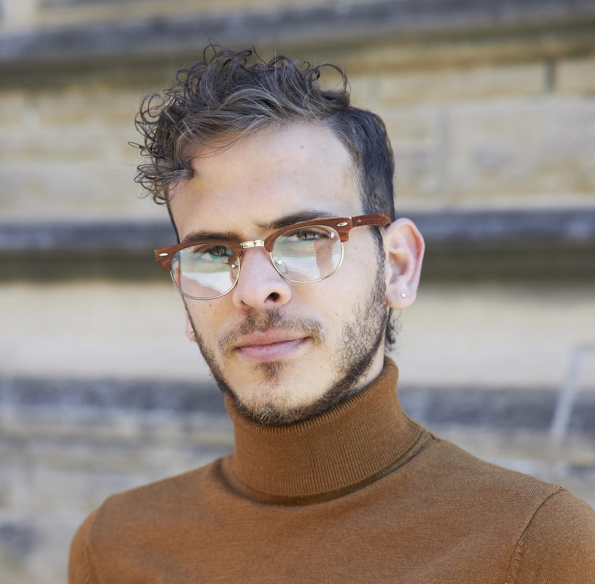 Close-up of a person with short, curly hair, wearing glasses and a brown turtleneck, with a blurred outdoor background.