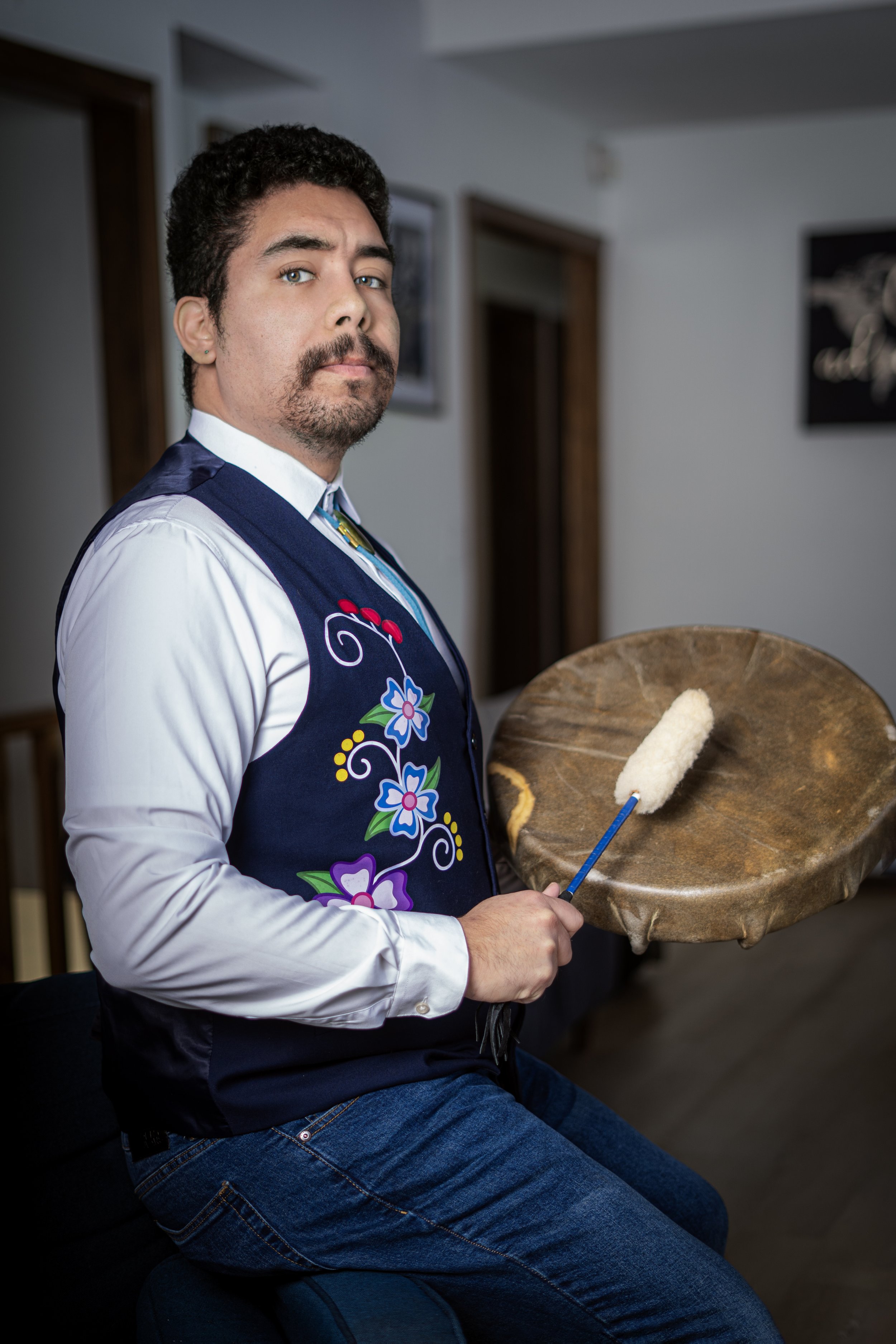 A man with dark curly hair, a mustache, and a goatee wearing a traditional embroidered vest over a white shirt, holding a wooden bowl with a paint roller in it, indoors with framed artwork on the wall.