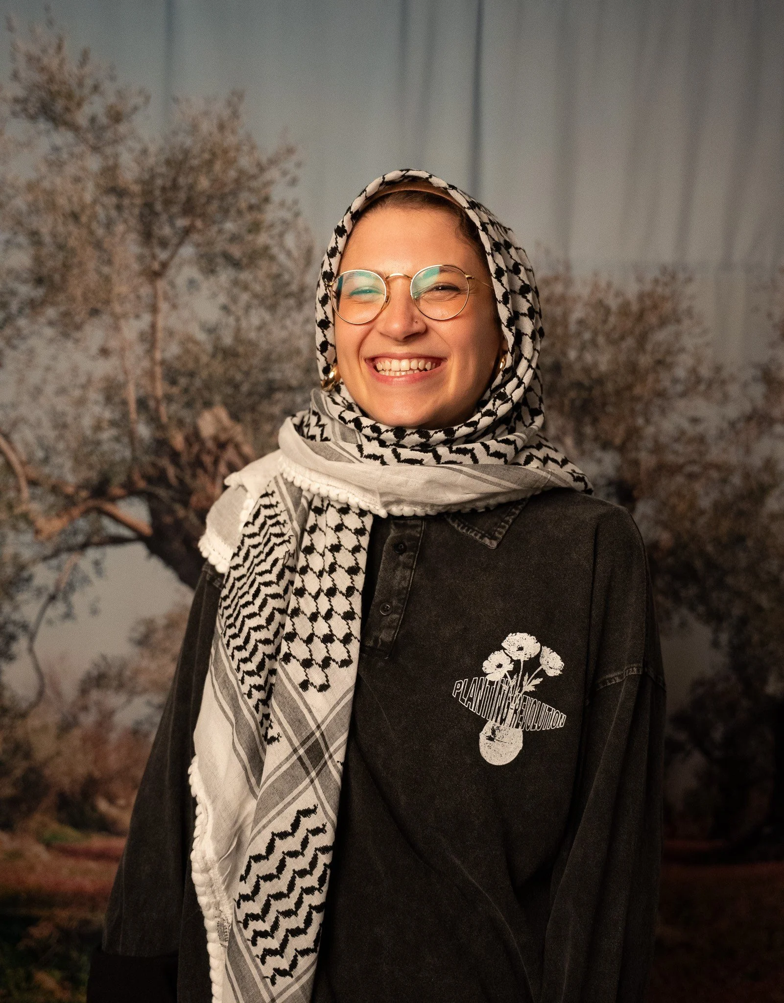 A young woman wearing glasses and a patterned headscarf smiling broadly. She is dressed in a black jacket with white floral logo and text, standing outdoors with trees in the background.