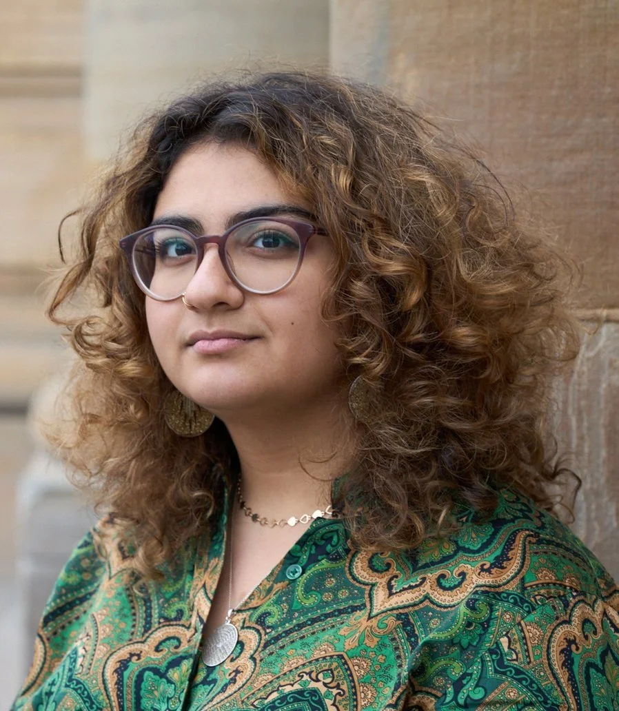 A woman with curly brown hair, wearing glasses, earrings, a necklace, and a green paisley patterned top, standing outdoors against a brick and sandstone background.