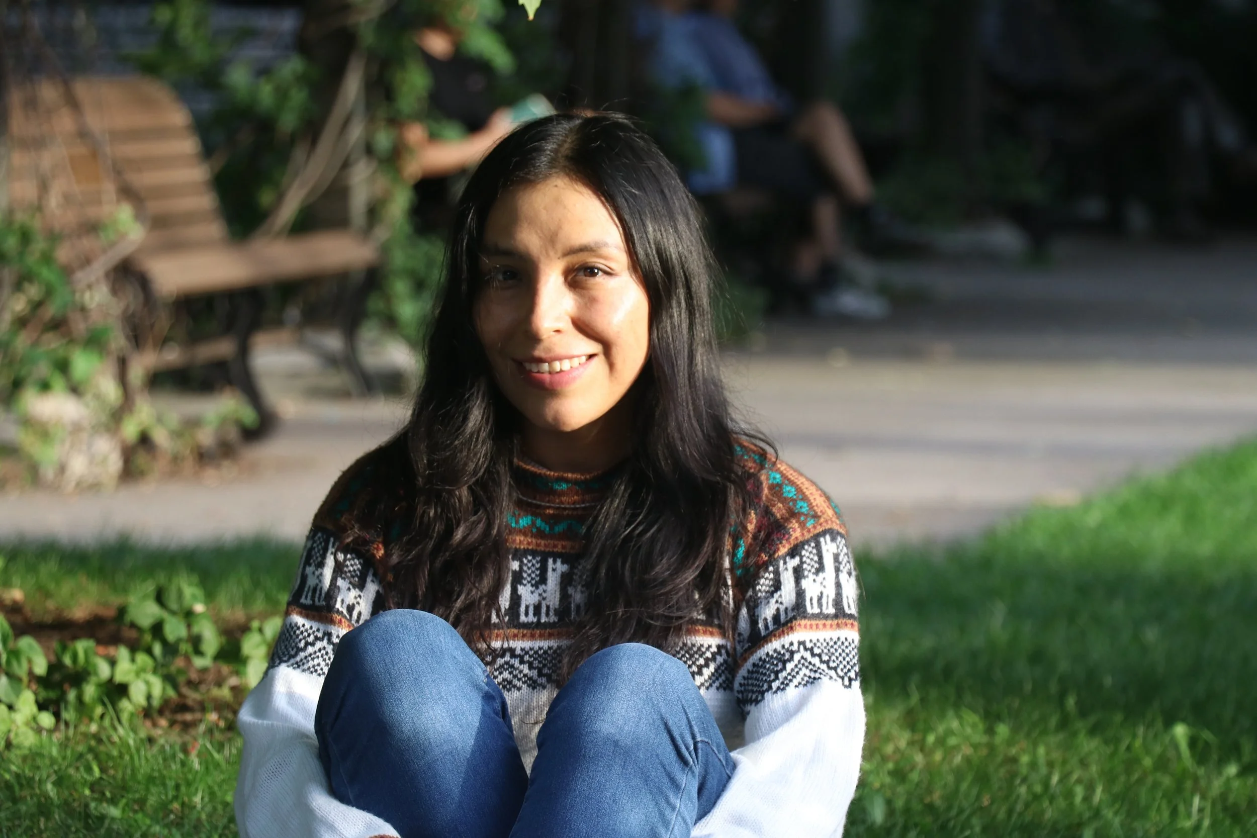 A smiling woman with dark hair and light skin tone sitting outdoors on the grass, wearing a patterned sweater and blue jeans, with blurred park benches and greenery in the background.