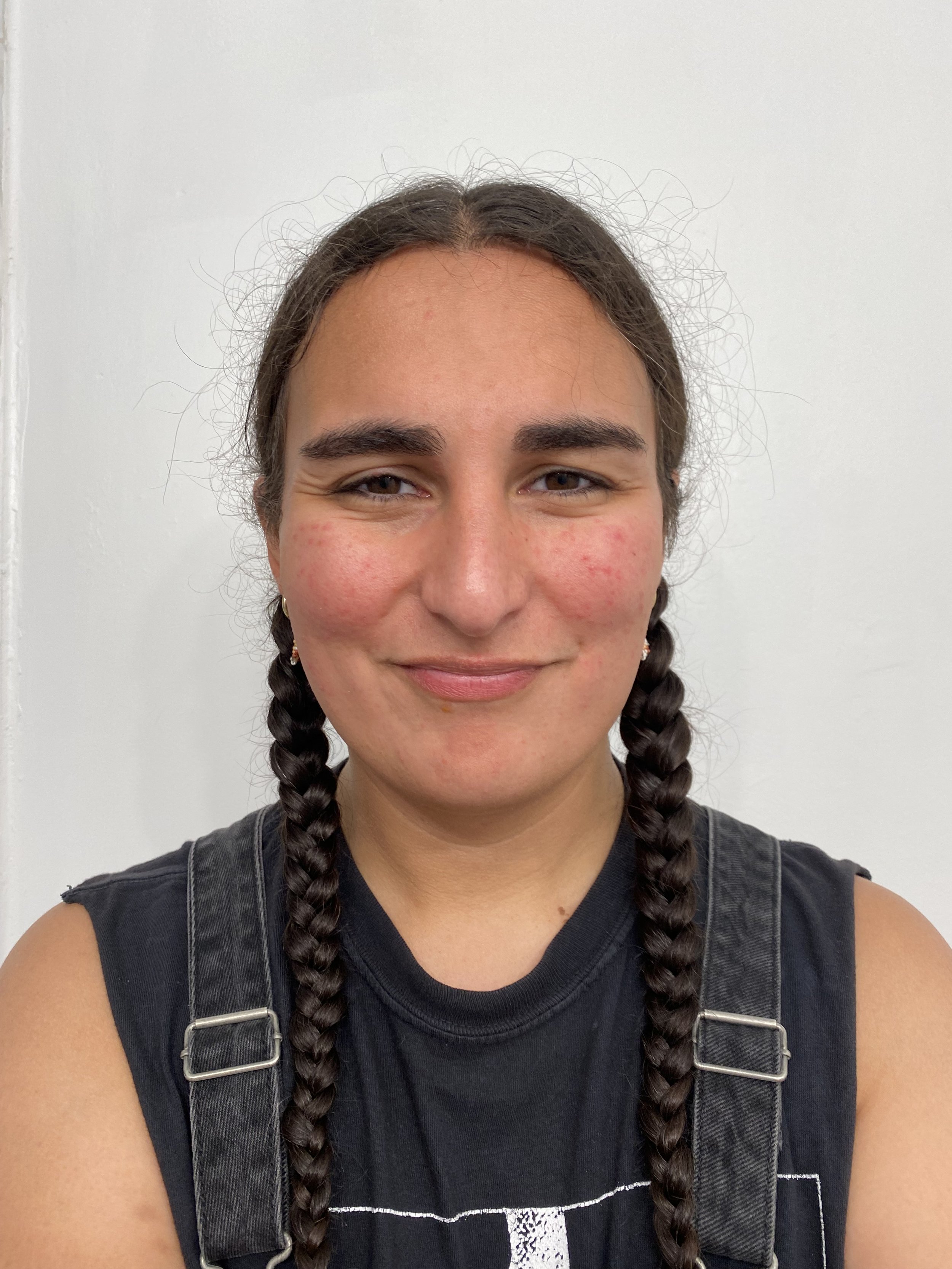 A young woman with braided hair, wearing a black sleeveless shirt and gray overalls, smiling in front of a plain white wall.