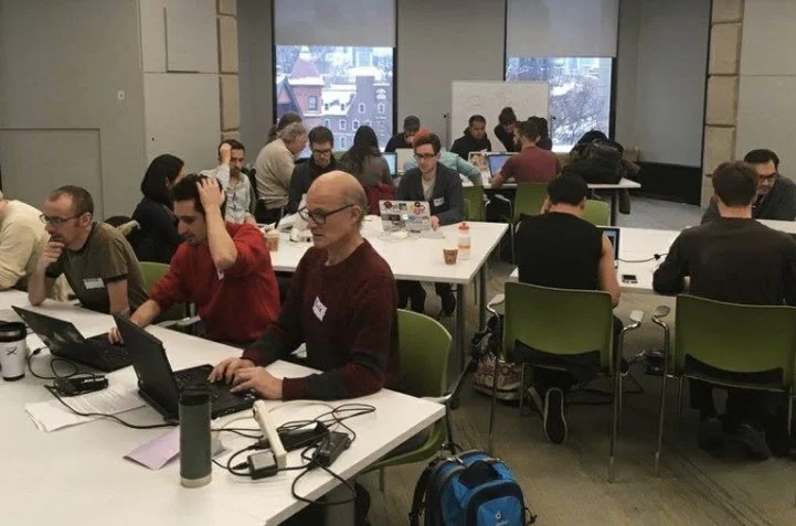 People working on laptops at tables in a well-lit office or conference room with large windows showing buildings outside.