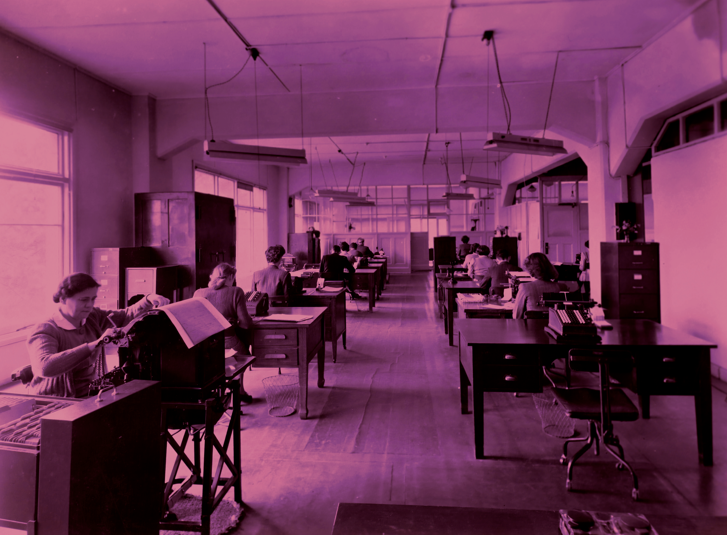 Classroom with students sitting at desks, studying, under pink lighting, with large windows and ceiling lights.
