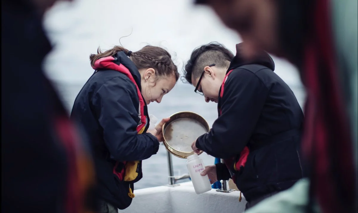 Two people on a boat, collecting water in a container, with other individuals partially visible around them.