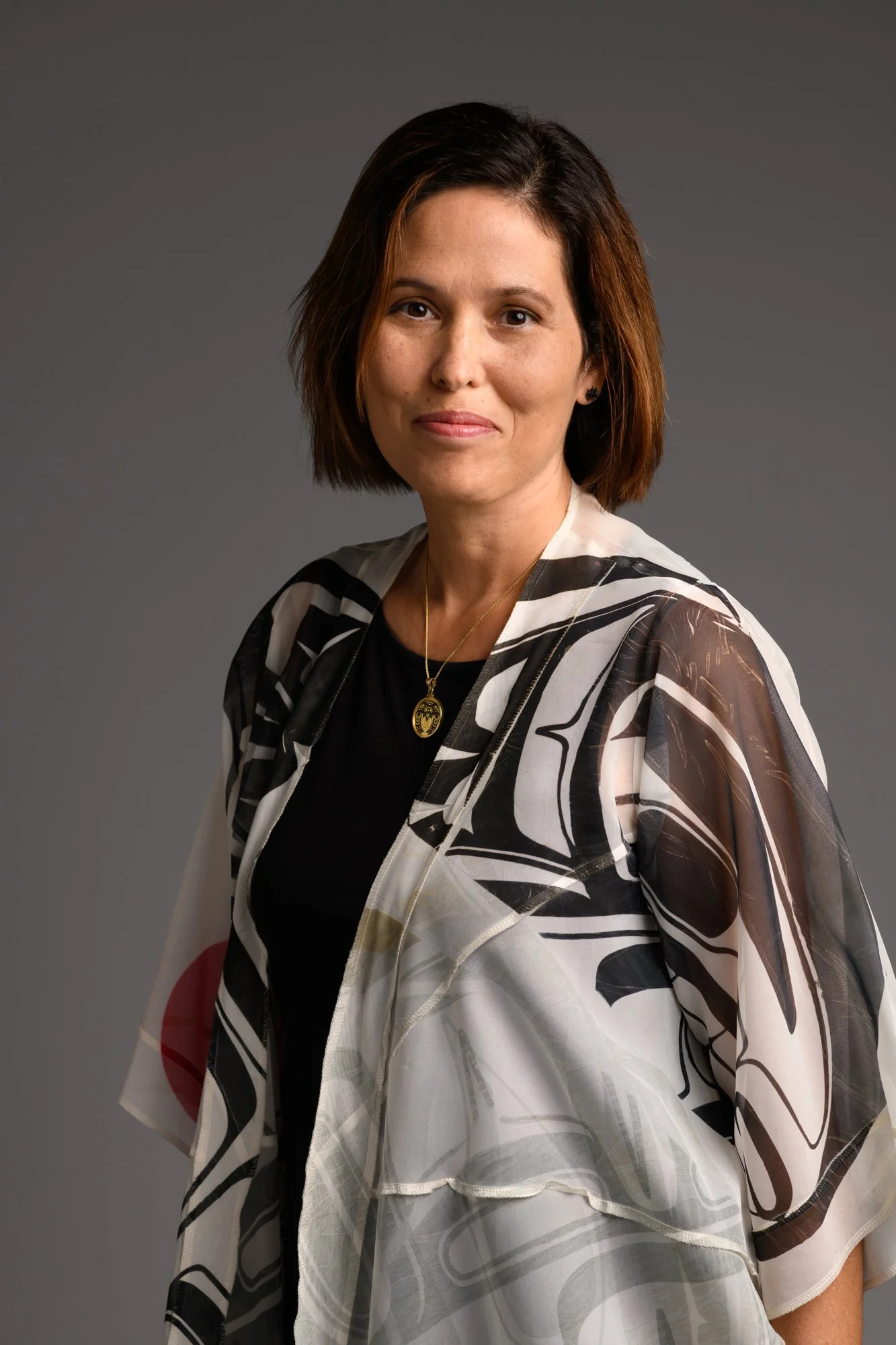 A woman with shoulder-length brown hair wearing a black top and a sheer, patterned white and black jacket, standing against a plain gray background.