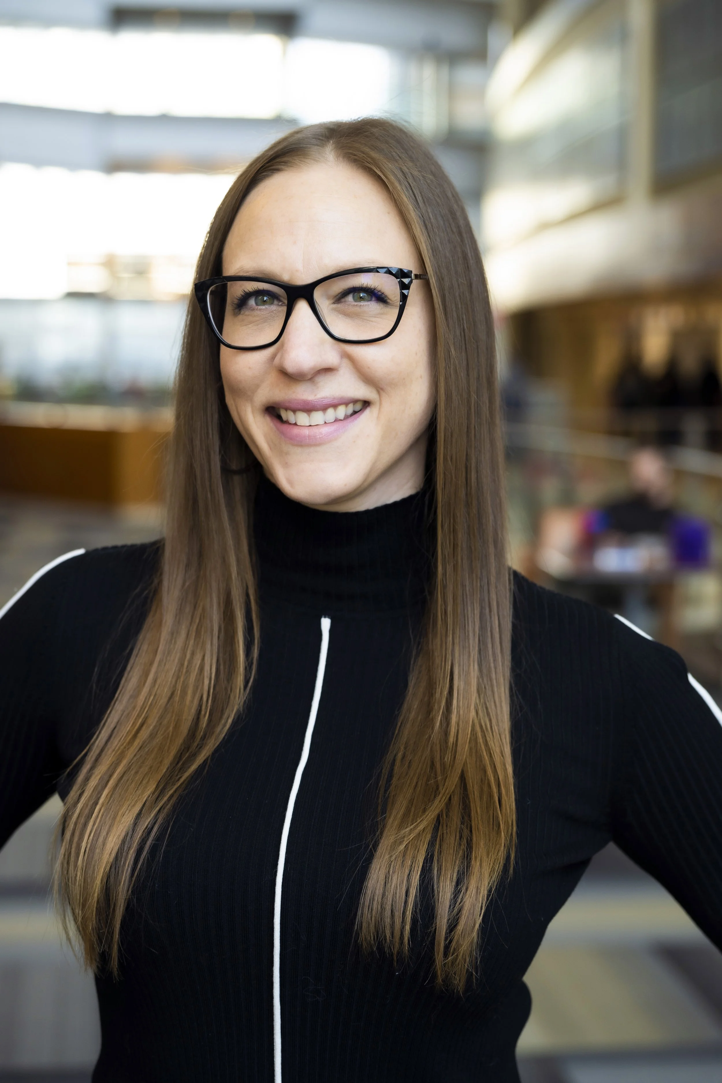 Close-up of a smiling woman with long brown hair wearing black glasses and a black zip-up jacket in an indoor setting.