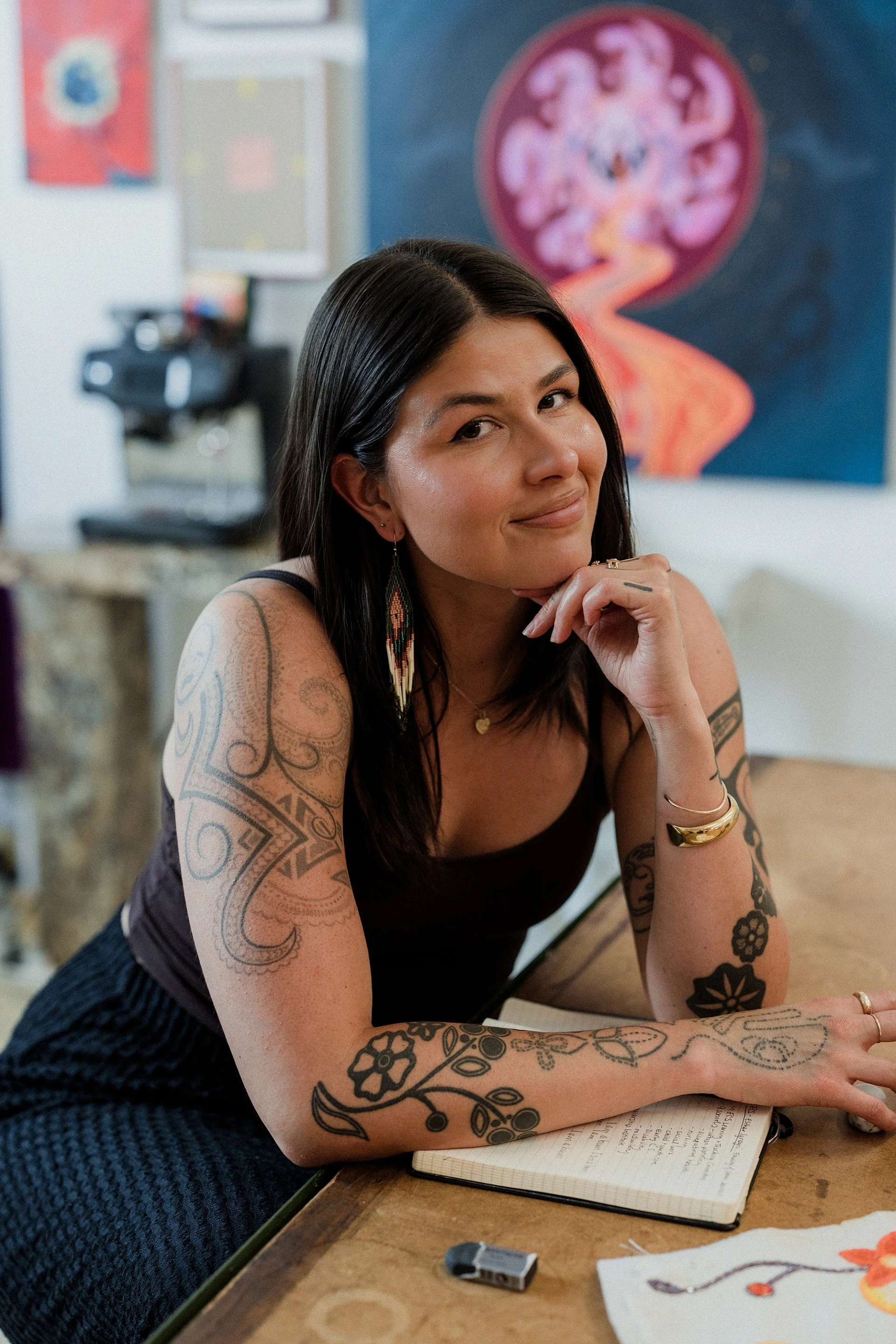 A woman with long black hair and tattoos on her arms rests her chin on her hand while sitting at a wooden table with a notebook, pen, and other art supplies. Behind her, there is a colorful painting on the wall and a microscope.