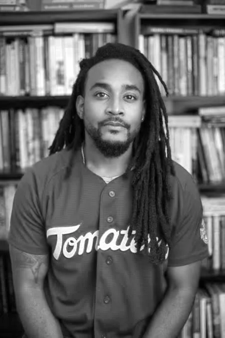 A man with dreadlocks and a beard wearing a shirt with the word 'Tomato' on it, standing in front of a bookshelf filled with books.
