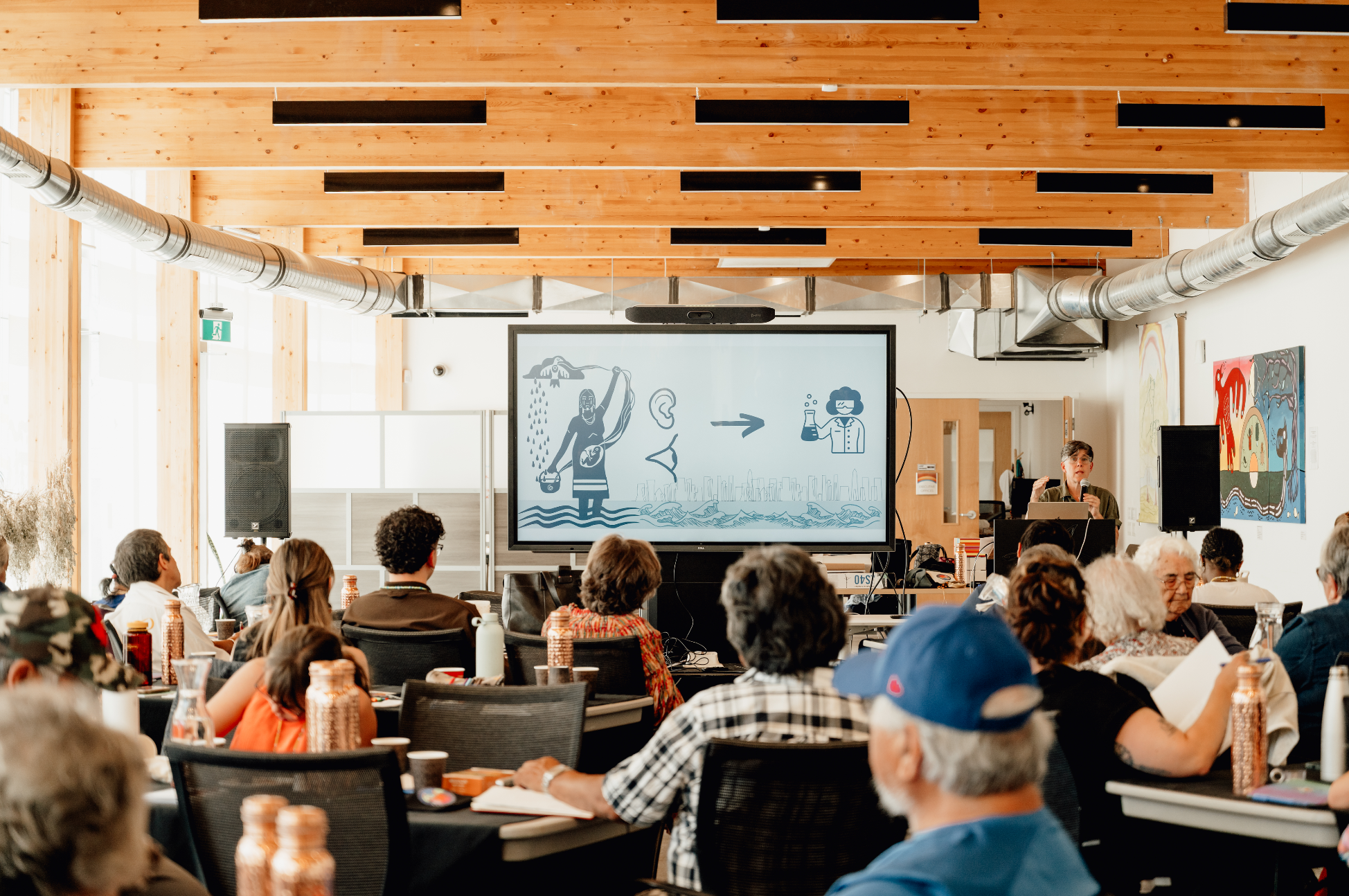 A conference room filled with people attending a presentation. There is a presenter at the front near a large screen displaying a diagram. The room has wooden ceiling beams and ductwork visible overhead.