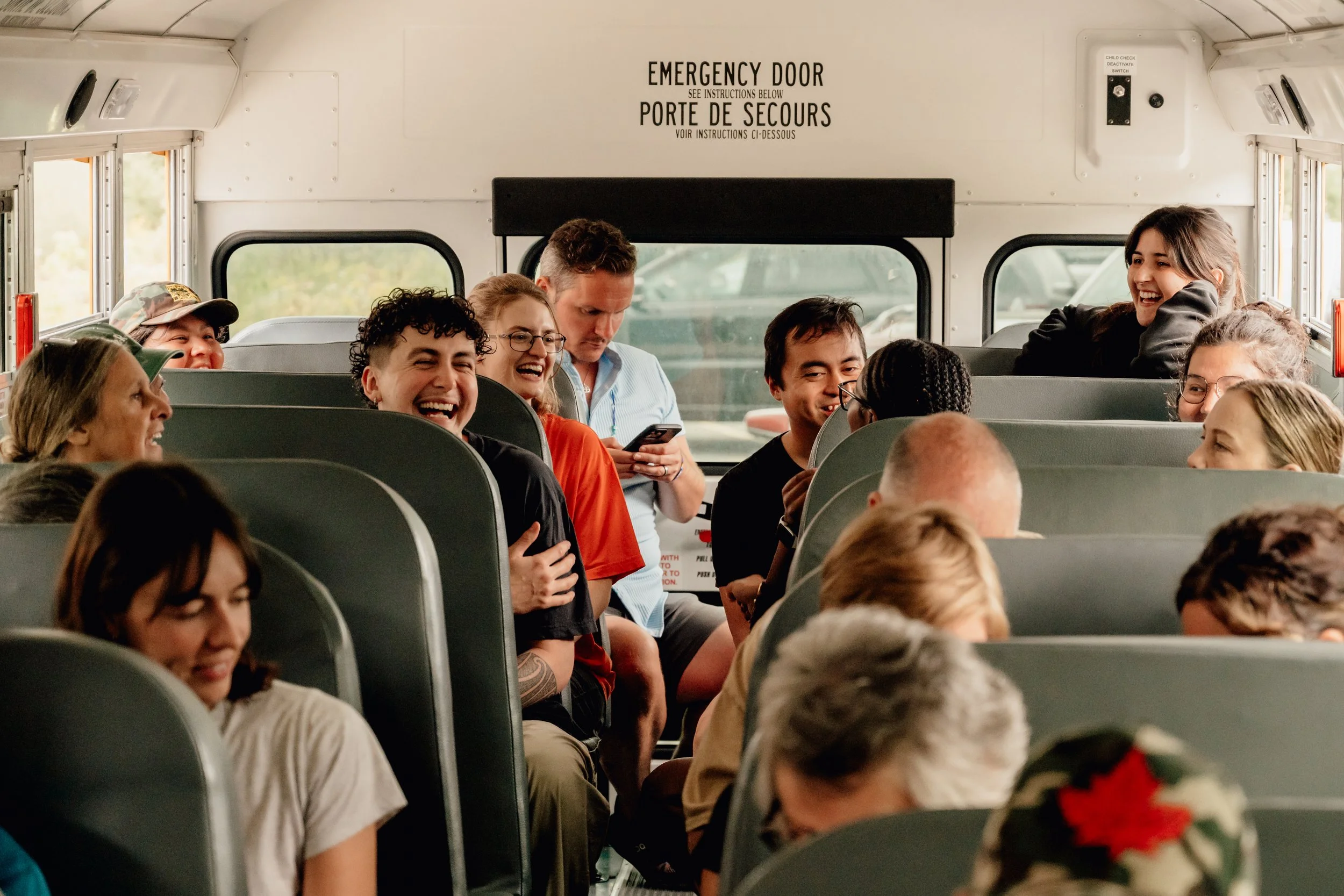 A group of people on a bus, some are smiling and laughing, one person is looking at their phone, and others are engaged in conversation, with windows showing outside scenery.