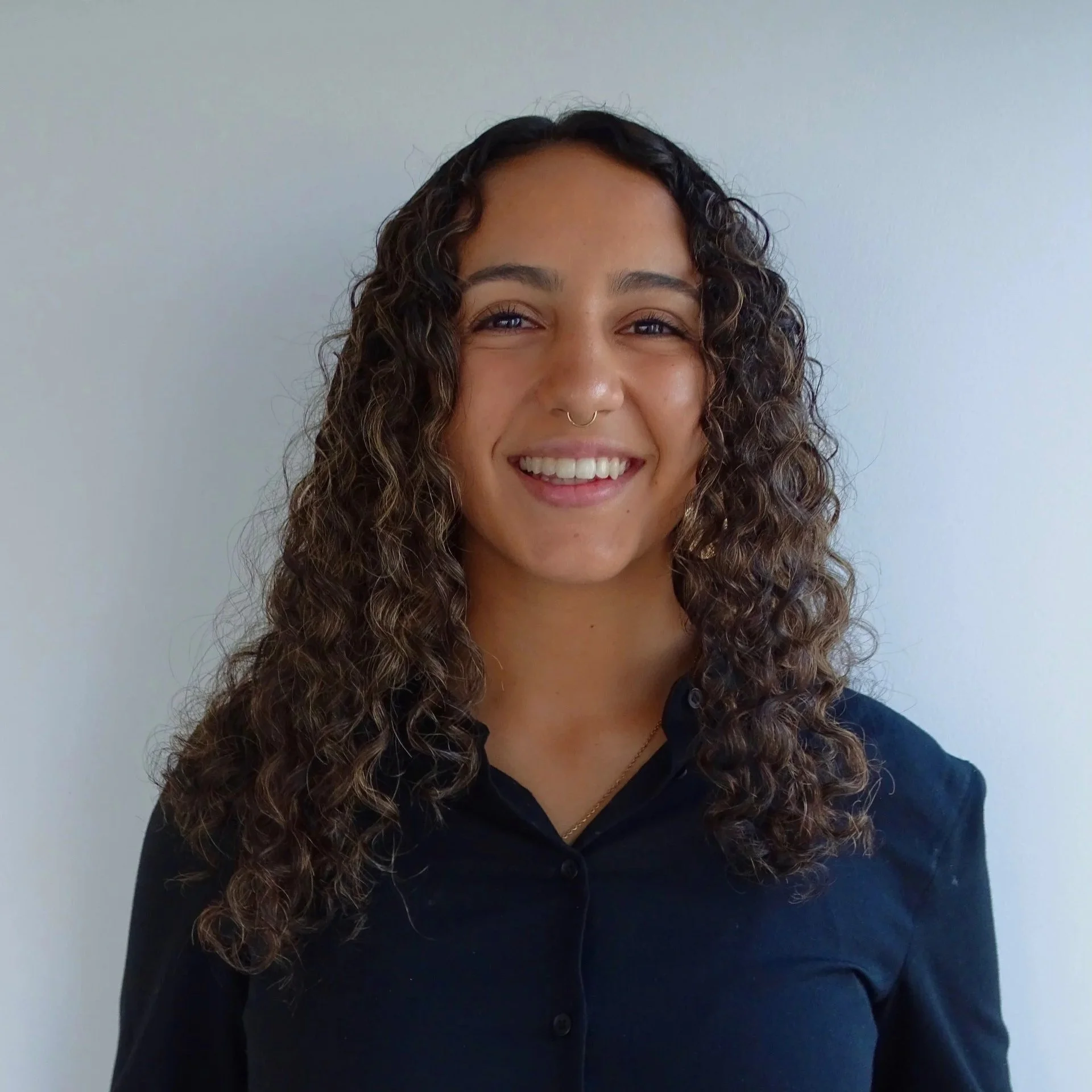 Close-up of a smiling woman with curly hair and a septum piercing, wearing a navy blue shirt, standing against a plain, light-colored wall.