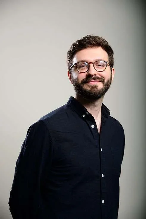 Portrait of a man with glasses and a beard, wearing a black button-up shirt, standing against a neutral background.
