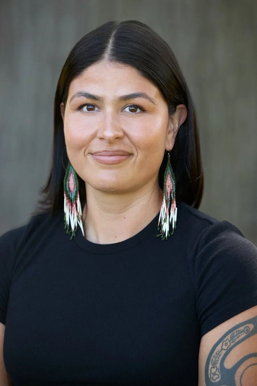 A woman with straight black hair wearing large beaded earrings and a black shirt, standing outdoors with a blurred natural background.