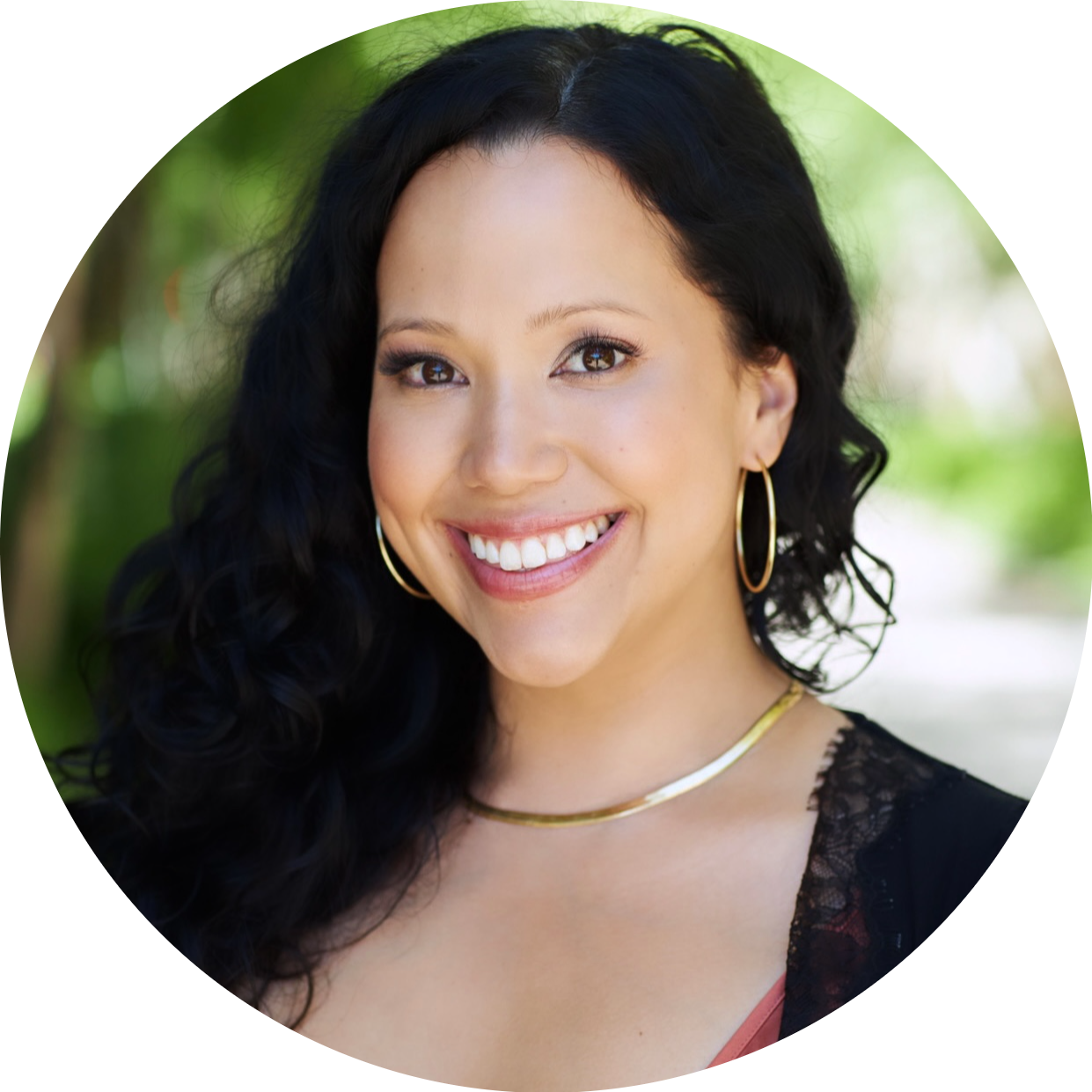 Close-up of a smiling woman with black wavy hair, wearing hoop earrings, a gold necklace, and a black lace top, outdoors with a blurred green background.