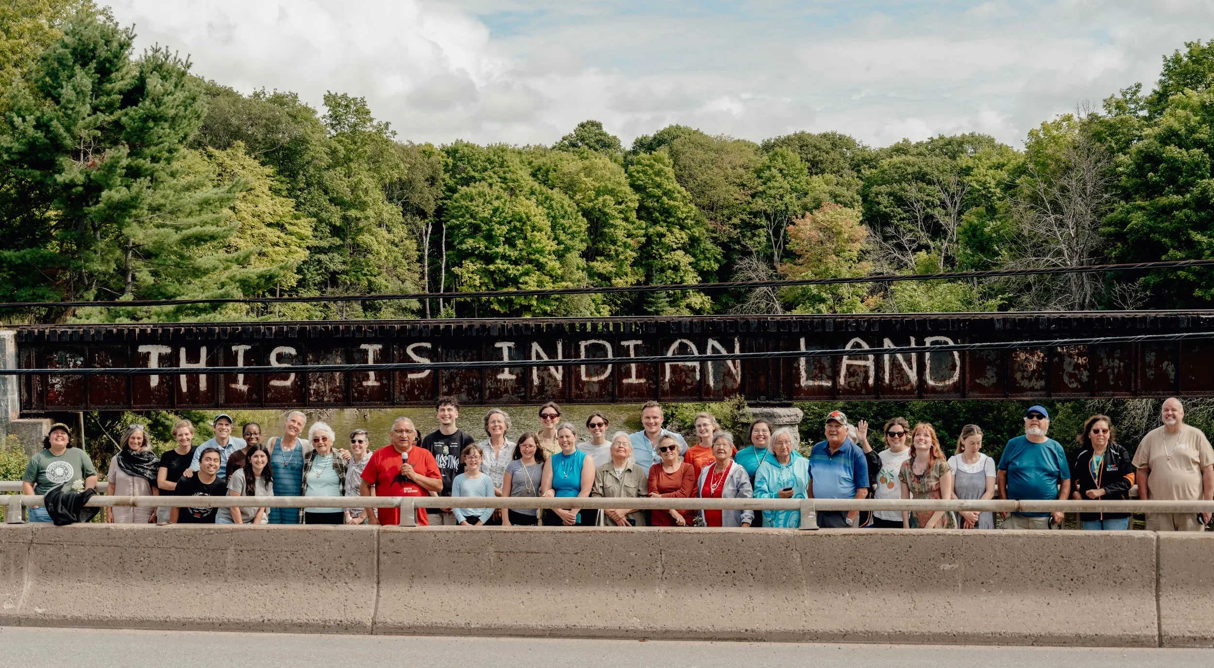 A large group of people standing on a bridge in front of a sign that reads 'This is Indiana.' The group is diverse in age and ethnicity, smiling for the photo, with lush green trees and a cloudy sky in the background.