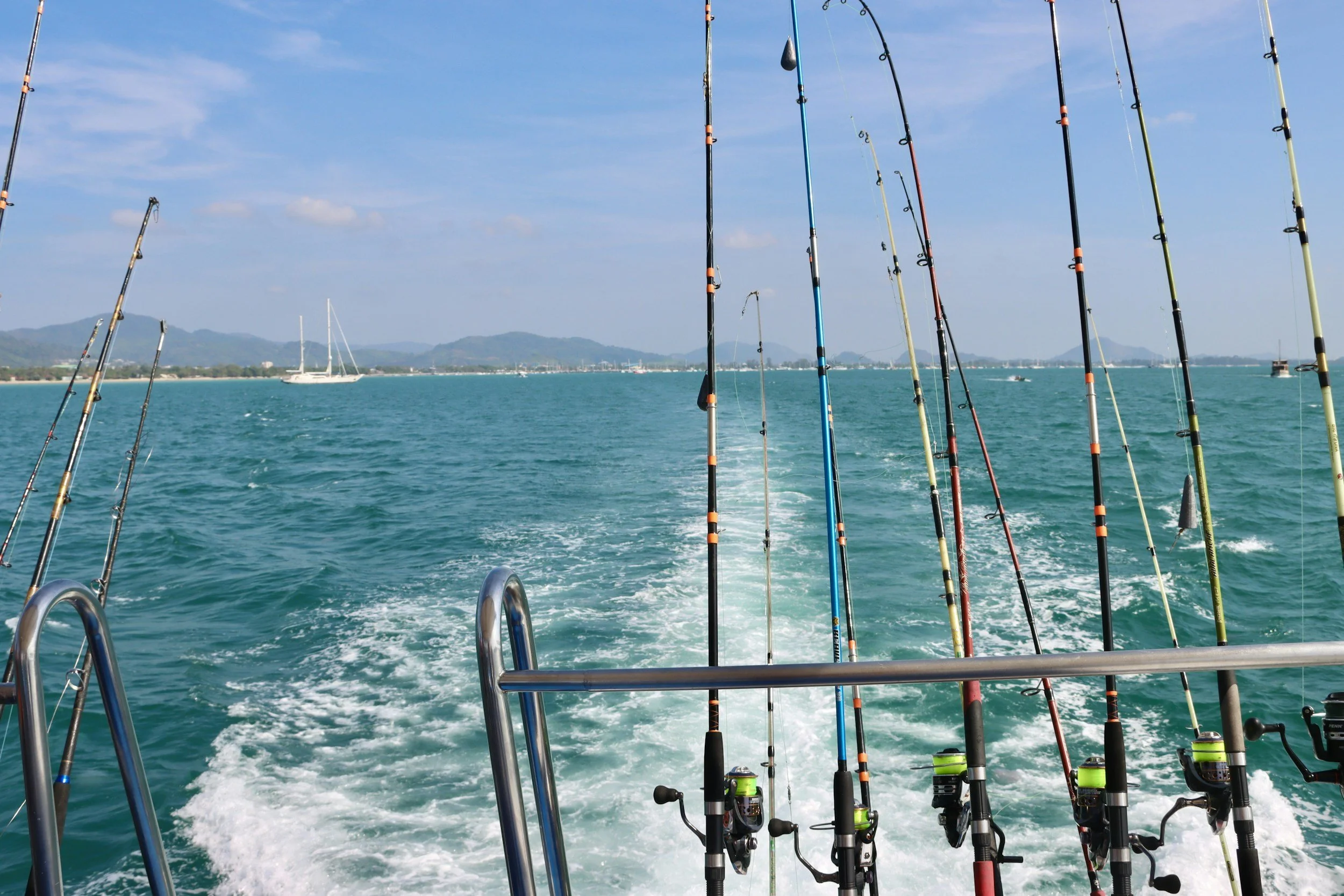 View from a boat with several fishing rods extending over the water, a distant sailboat, and mountains in the background under a partly cloudy sky.