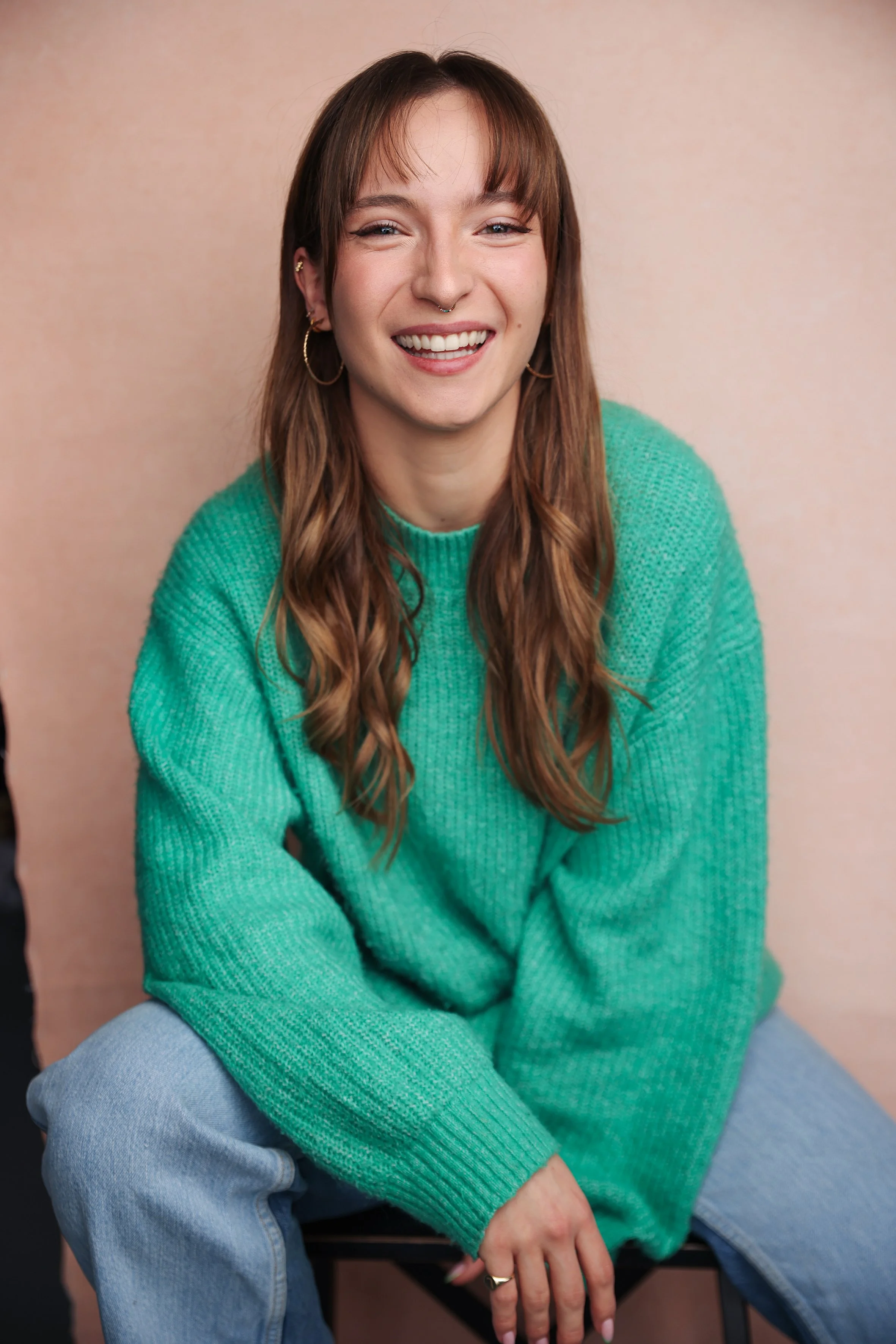 A young woman with brown hair, bright eyes, and a big smile wearing a green sweater and light blue jeans, sitting against a beige background.