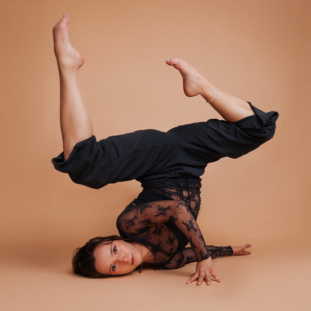 A woman performing a headstand with her legs bent, wearing a black lace top and black pants against a beige background.
