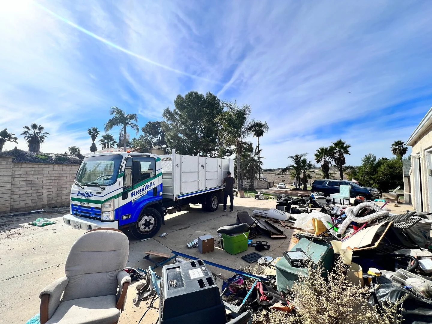 A cluttered driveway with a blue and white Republic truck, a black SUV, and various discarded items including furniture, pipes, and boxes. A person is standing near the truck under a sunny sky with scattered clouds and palm trees in the background.