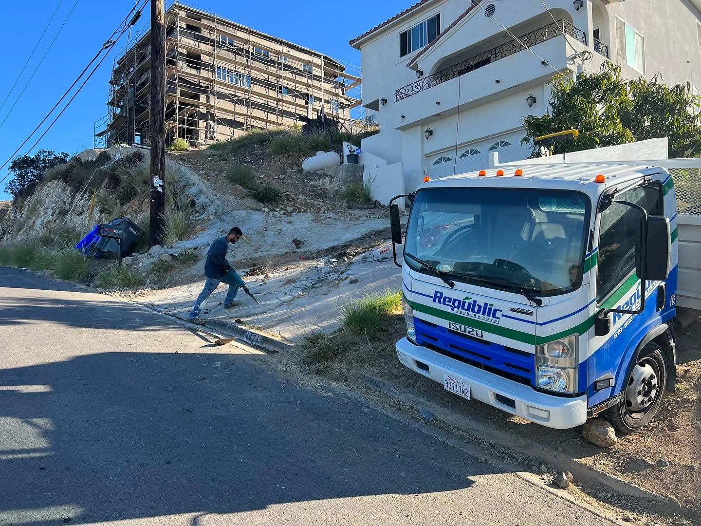 A worker using a tool to fix a damaged sidewalk on a hilly residential street with a truck nearby. There are houses and a construction site in the background.