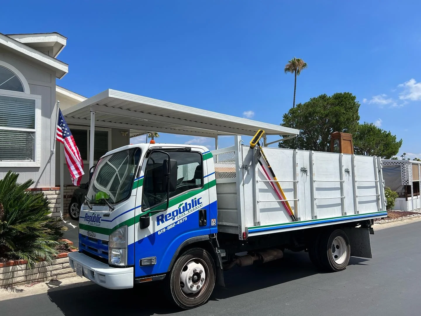A blue and white Republic truck parked in a residential area with palm trees, a house with a porch and an American flag, under a clear blue sky.