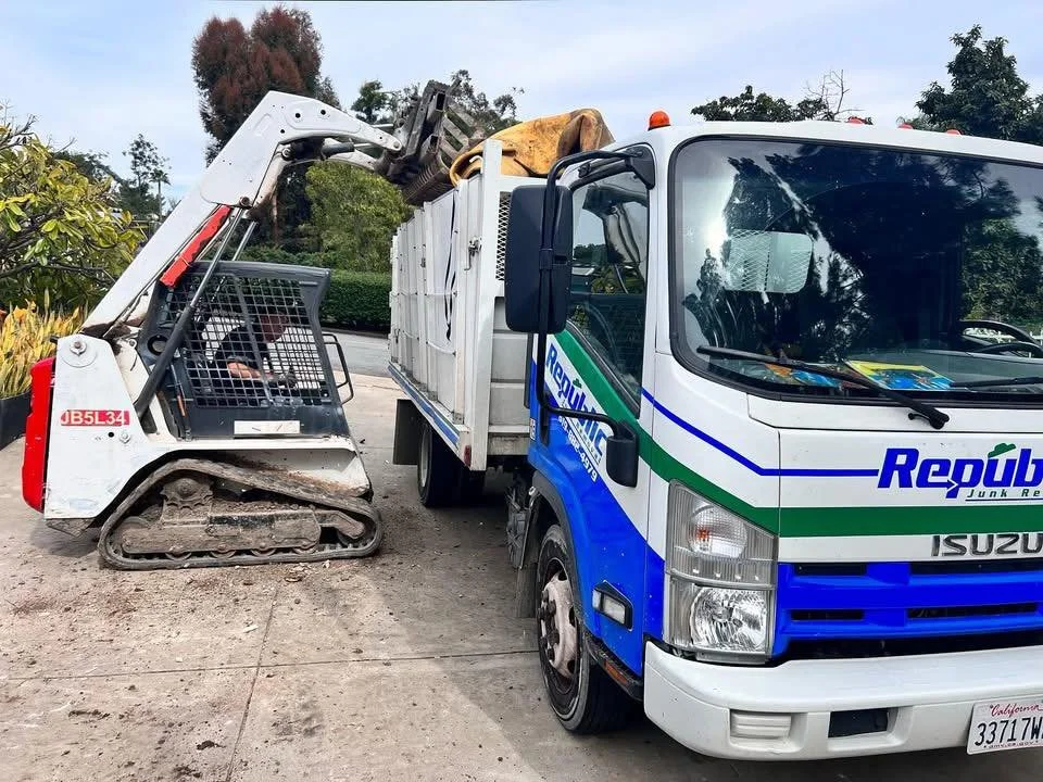 A skid-steer loader and a trash truck are positioned close to each other on a sidewalk, with the loader's arm extended toward the truck.