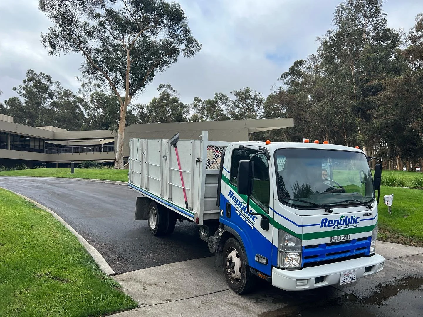 A white and blue garbage truck is parked on a paved driveway next to a grassy area with trees and a modern building in the background.