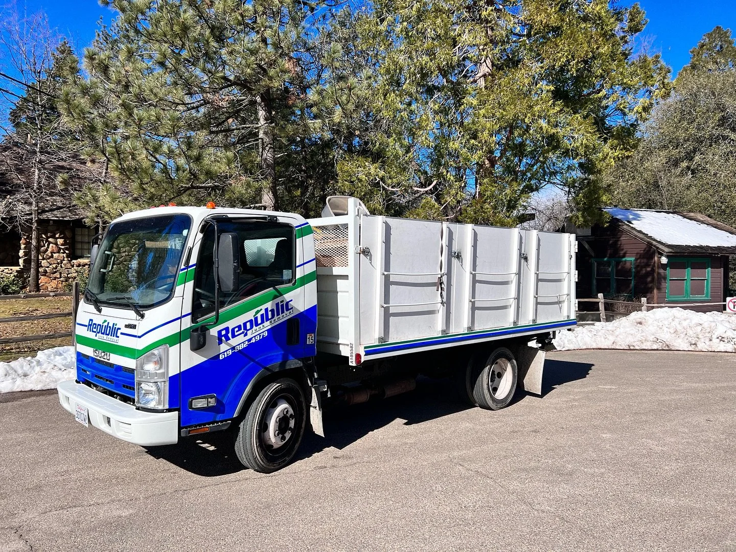 A blue and white Republic public works truck parked on a street with snow on the ground and trees in the background.