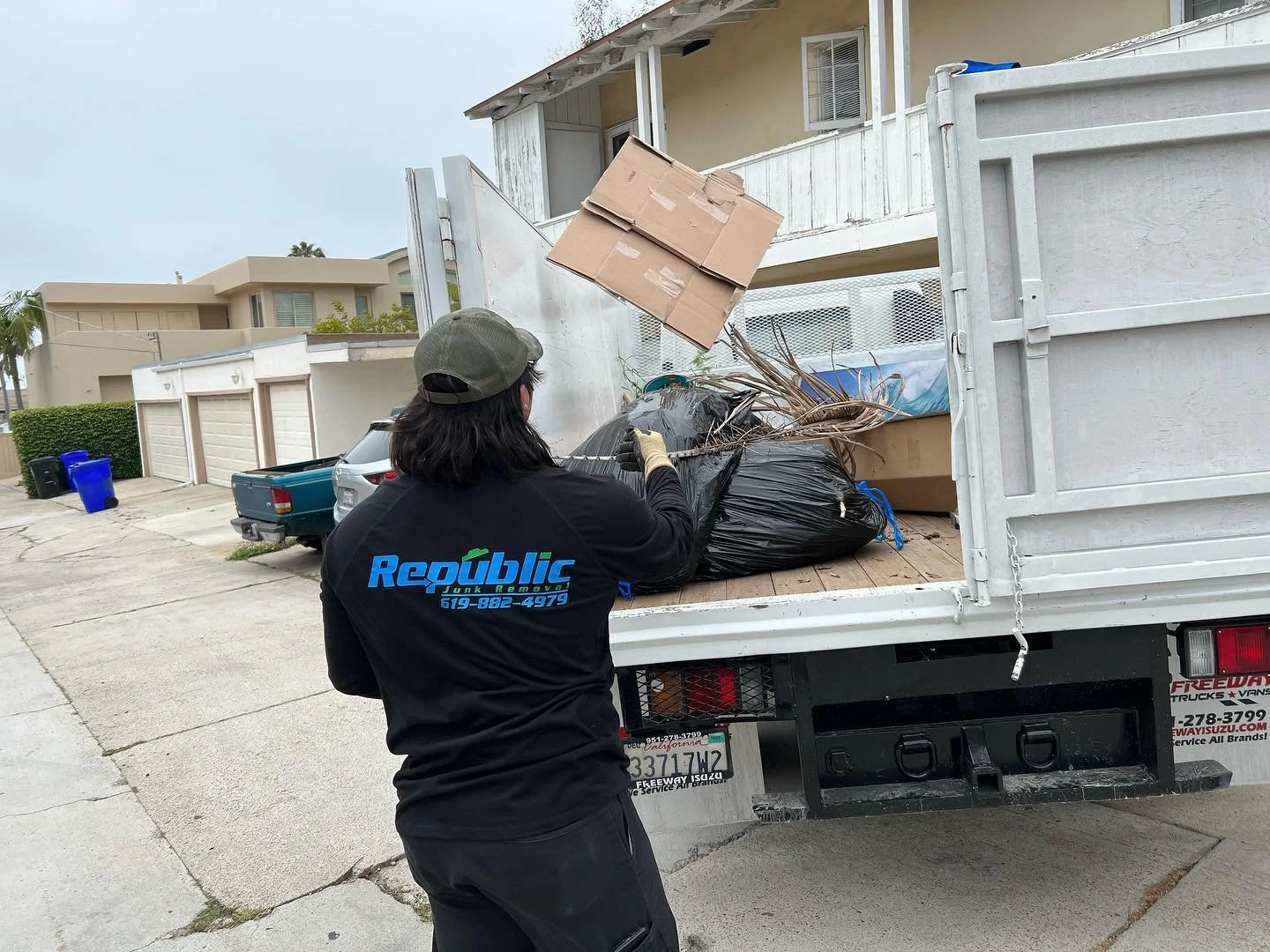 A man in a black shirt and baseball cap loading cardboard boxes and household trash into a truck, labeled 'Republic' with a phone number, parked on a residential street.
